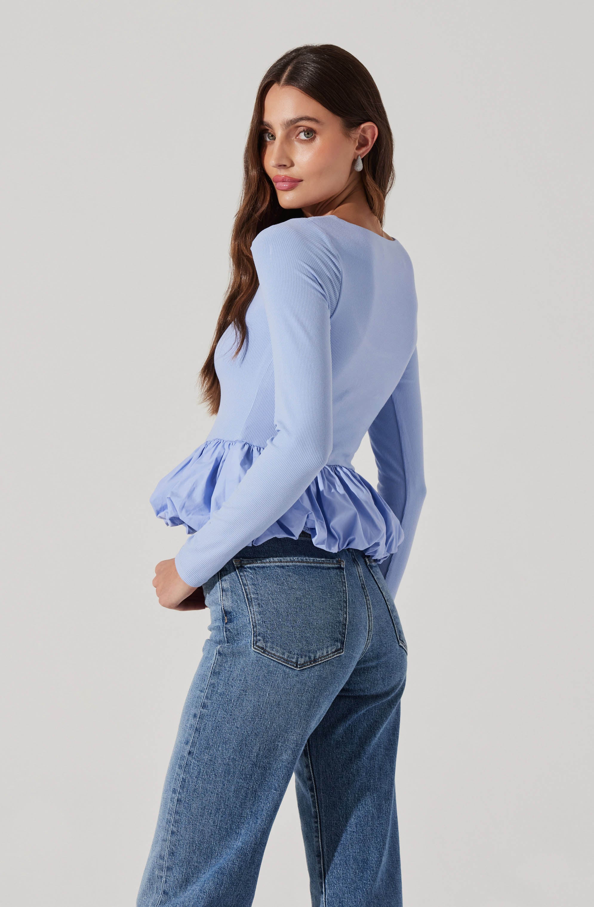 A woman is wearing a light blue long-sleeve top with a ruffled peplum hem and a pair of classic blue jeans.