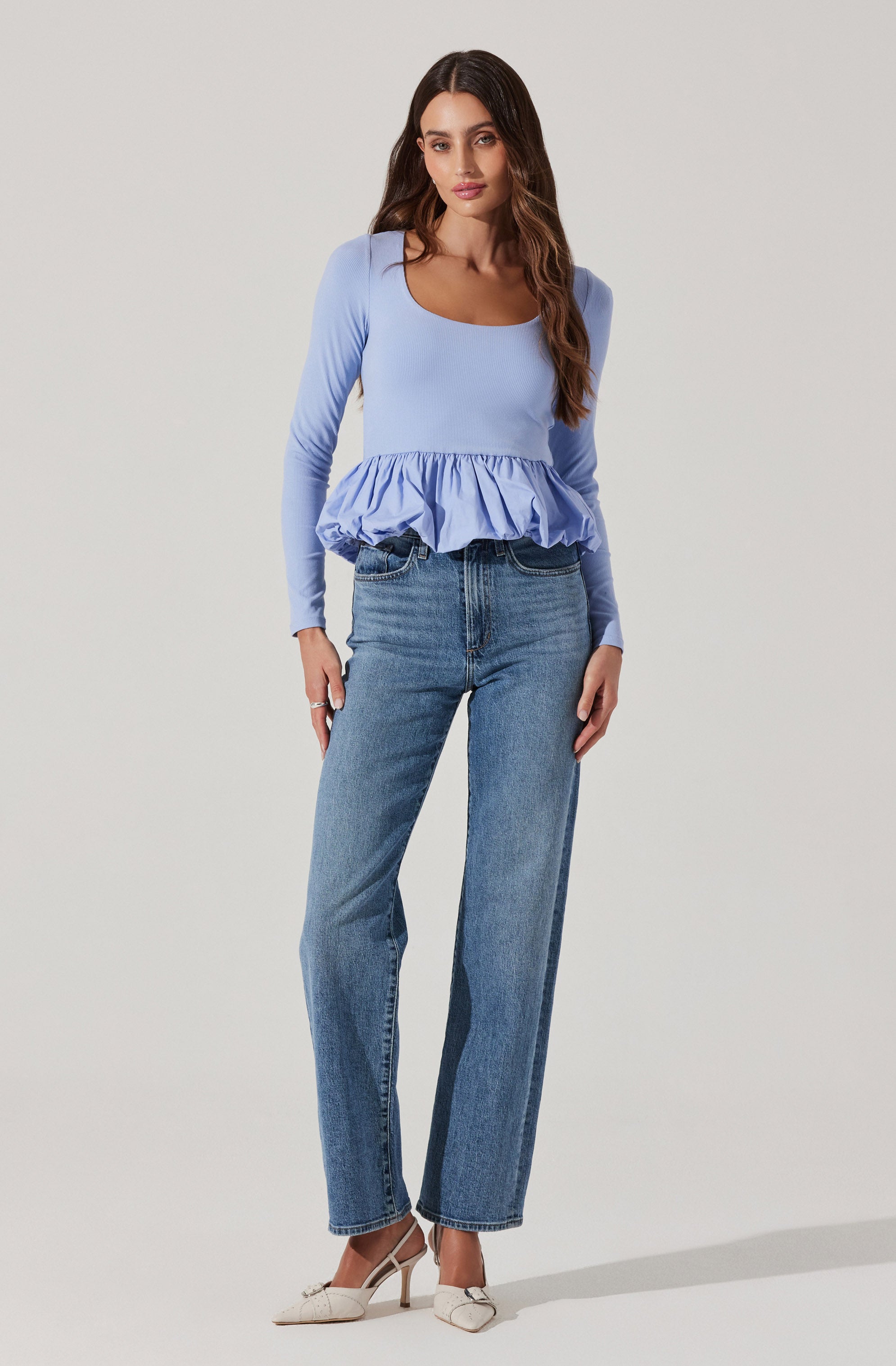 A woman is wearing a light blue long-sleeved top with a square neckline and a ruffled hem detail. She has paired it with high-waisted, straight-leg blue jeans and white pointed-toe heels with ankle straps.