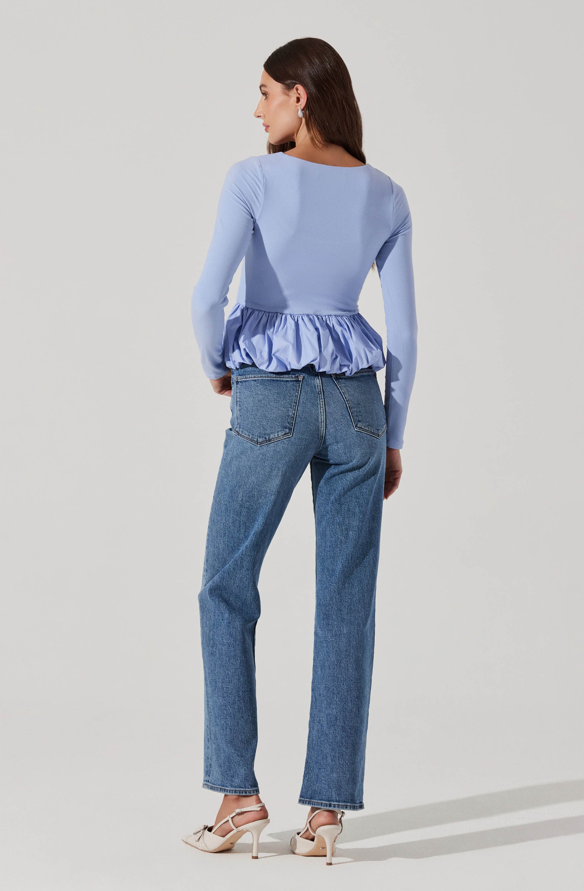 A woman is wearing a light blue long-sleeve top with a ruffled, satin-like band around the waist. She is also wearing high-waisted, straight-leg blue jeans and beige strappy high-heeled sandals.