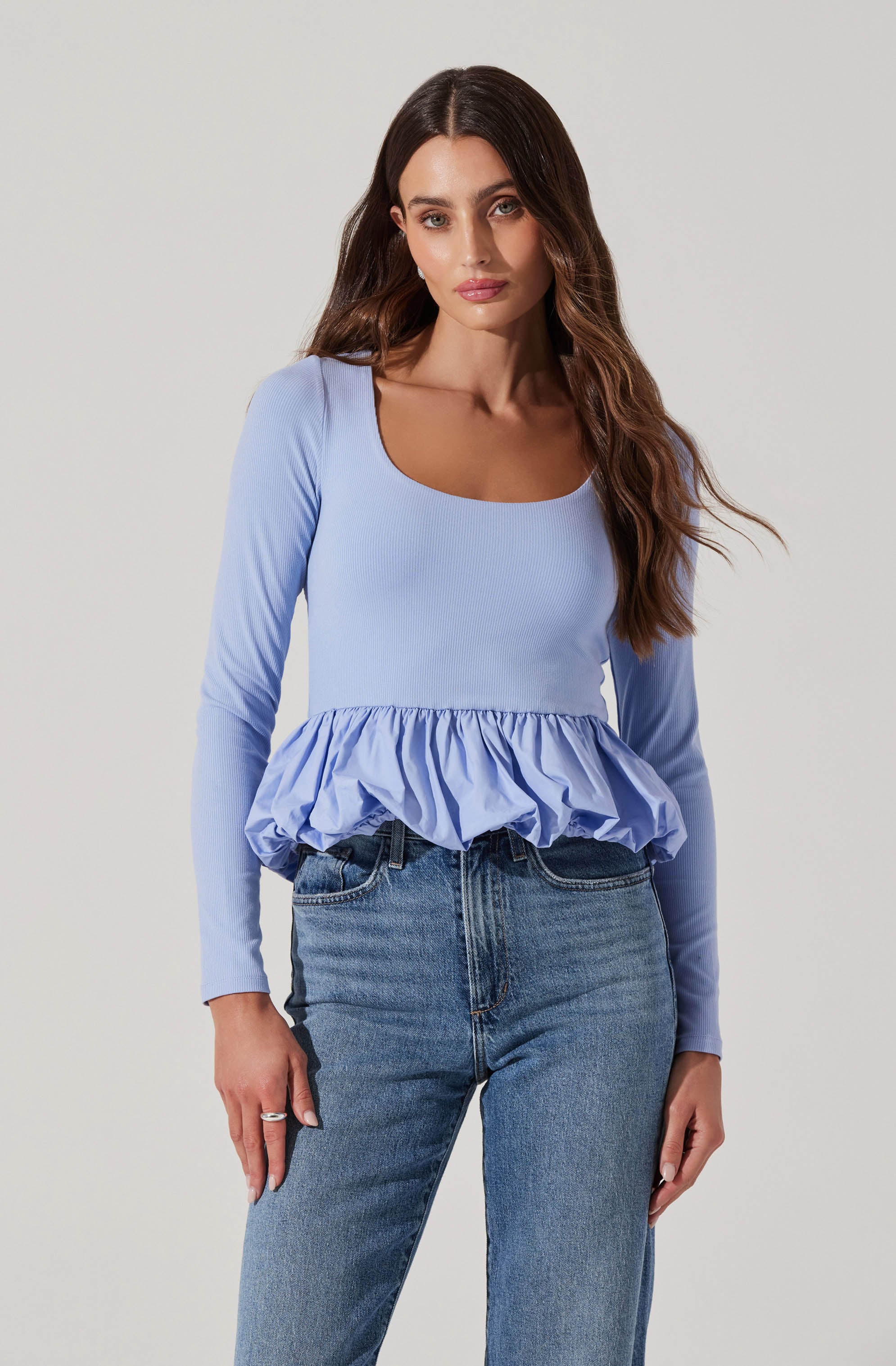 A woman is wearing a light blue, long-sleeved top with a round neckline. The top has a ruffled, gathered peplum detail at the waist. She is also wearing high-waisted blue jeans.
