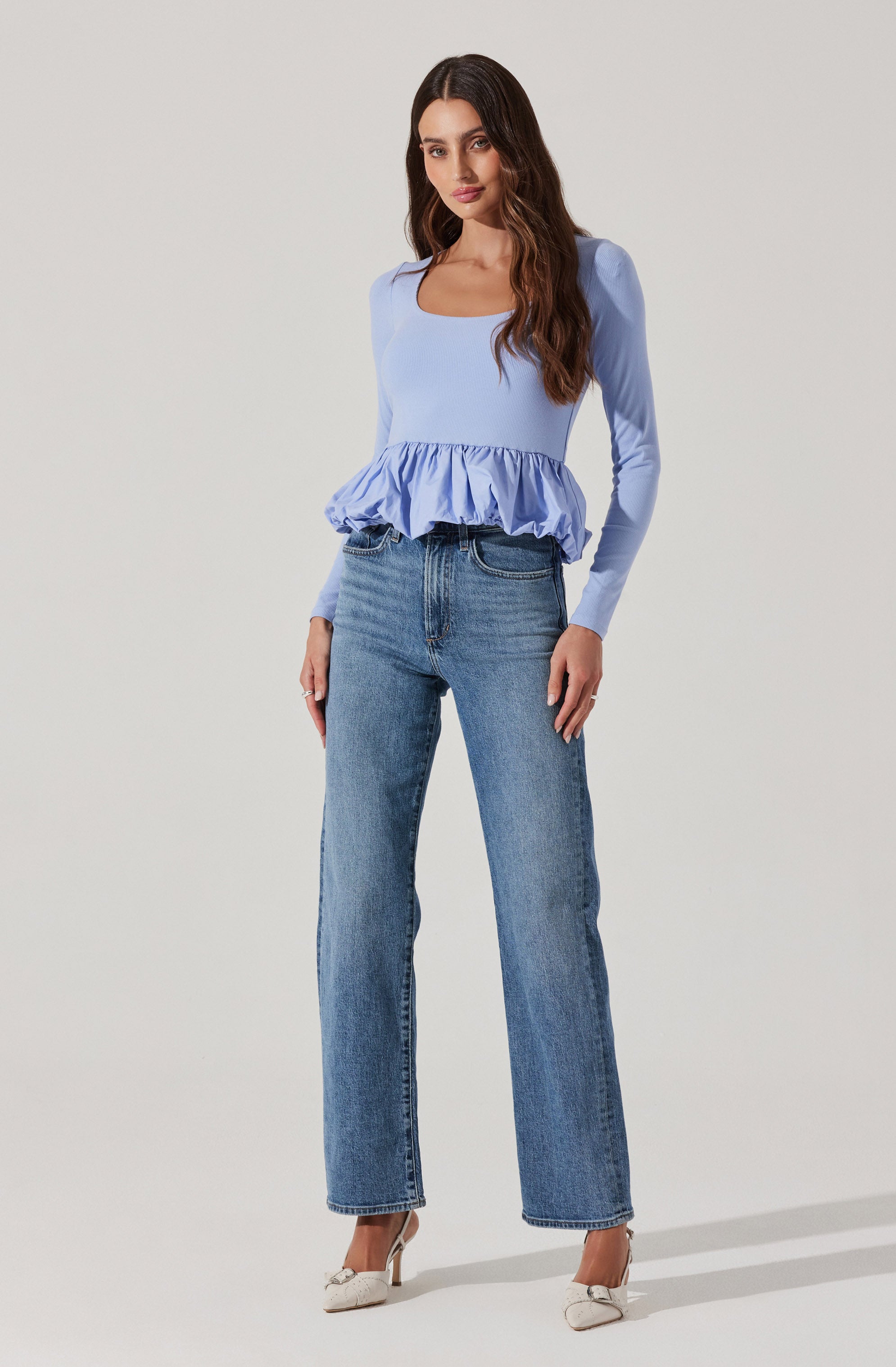A woman wearing a light blue long-sleeve top with a square neckline and a ruffled hem, paired with high-waisted, straight-leg blue jeans, and beige heels.