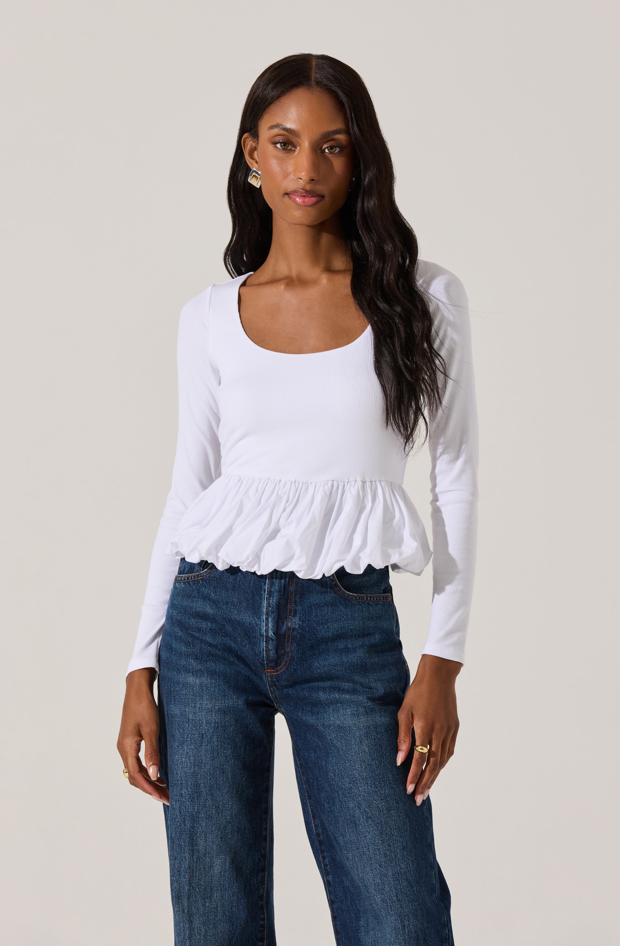 A woman is wearing a long-sleeved white blouse with a scoop neckline and a ruffled peplum hem, paired with high-waisted blue jeans.