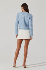 A woman is wearing a soft, light blue, textured long-sleeve sweater that appears cozy and slightly oversized. She pairs it with a high-waisted, short, white skirt that has a clean, simple design. She completes the outfit with beige high-heeled shoes with ankle straps.