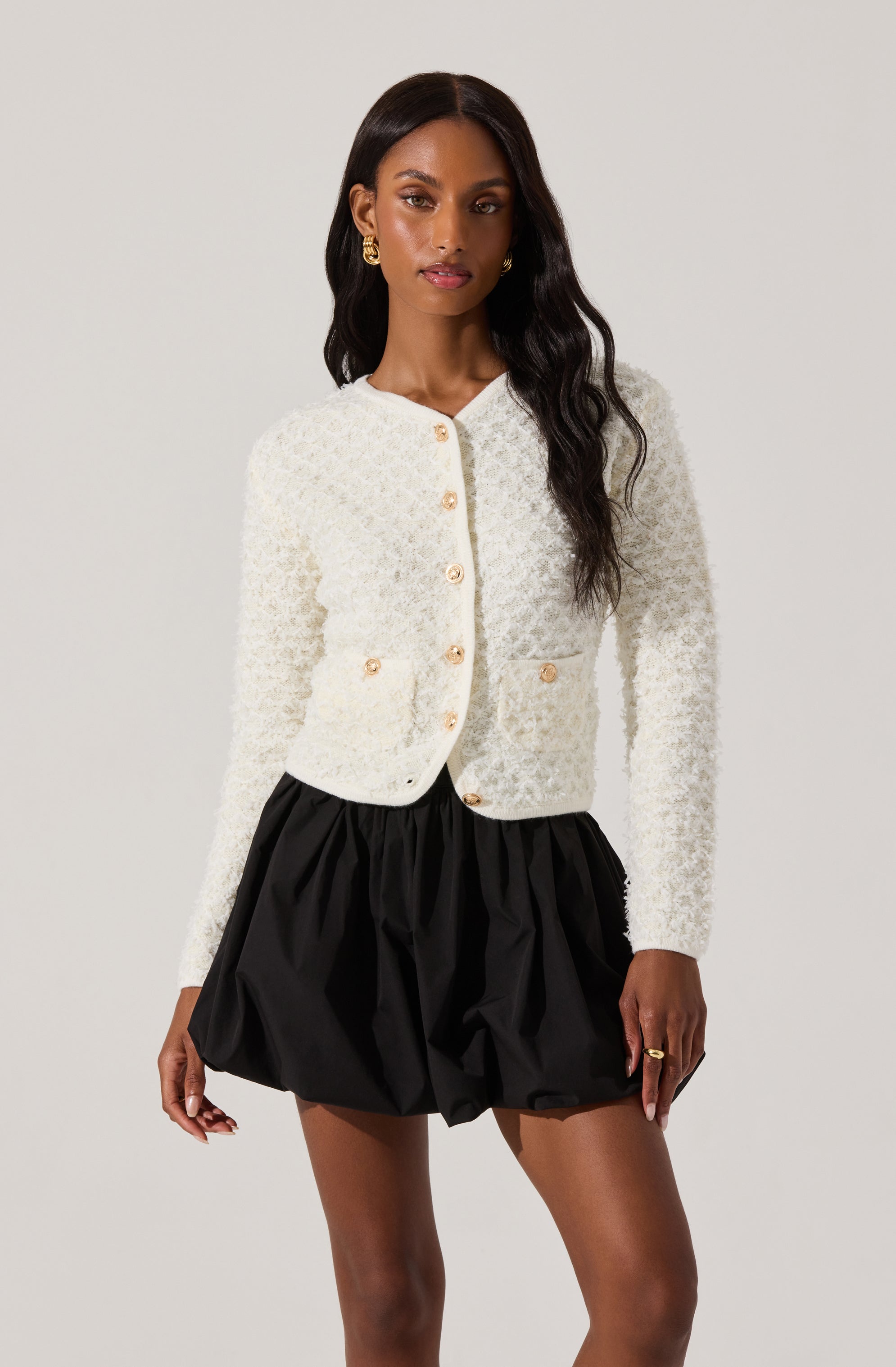 A woman is wearing a white textured, long-sleeved, button-up cardigan with gold buttons, paired with a black high-waisted, puffy, skirted shorts.