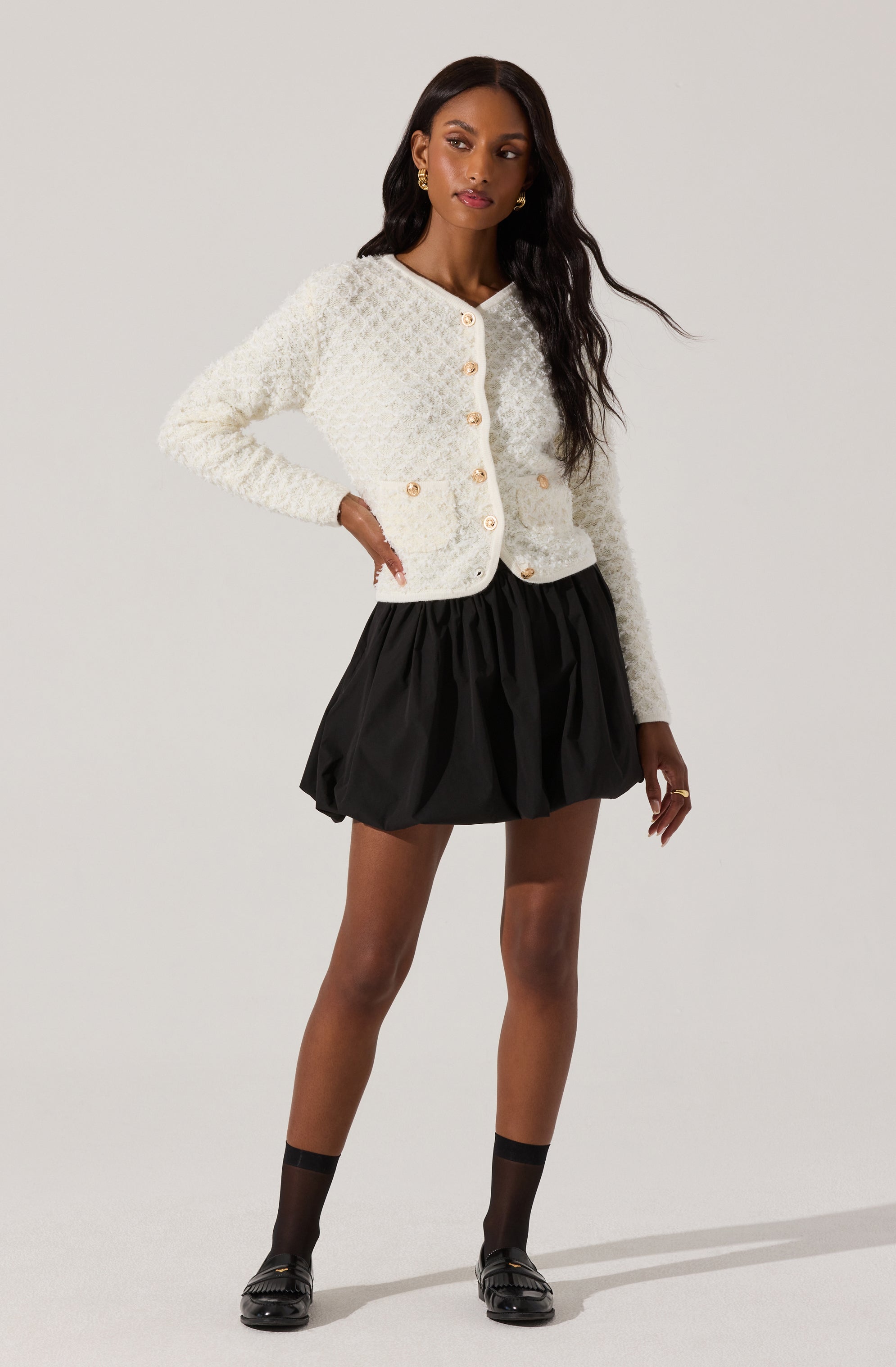 A woman is wearing a textured, long-sleeved, white knit cardigan with a rounded neckline and gold buttons down the front and on the pockets. She pairs it with a black, voluminous, above-the-knee skirt that has a gathered, bubble-like silhouette. She completes her outfit with black socks and black loafers.