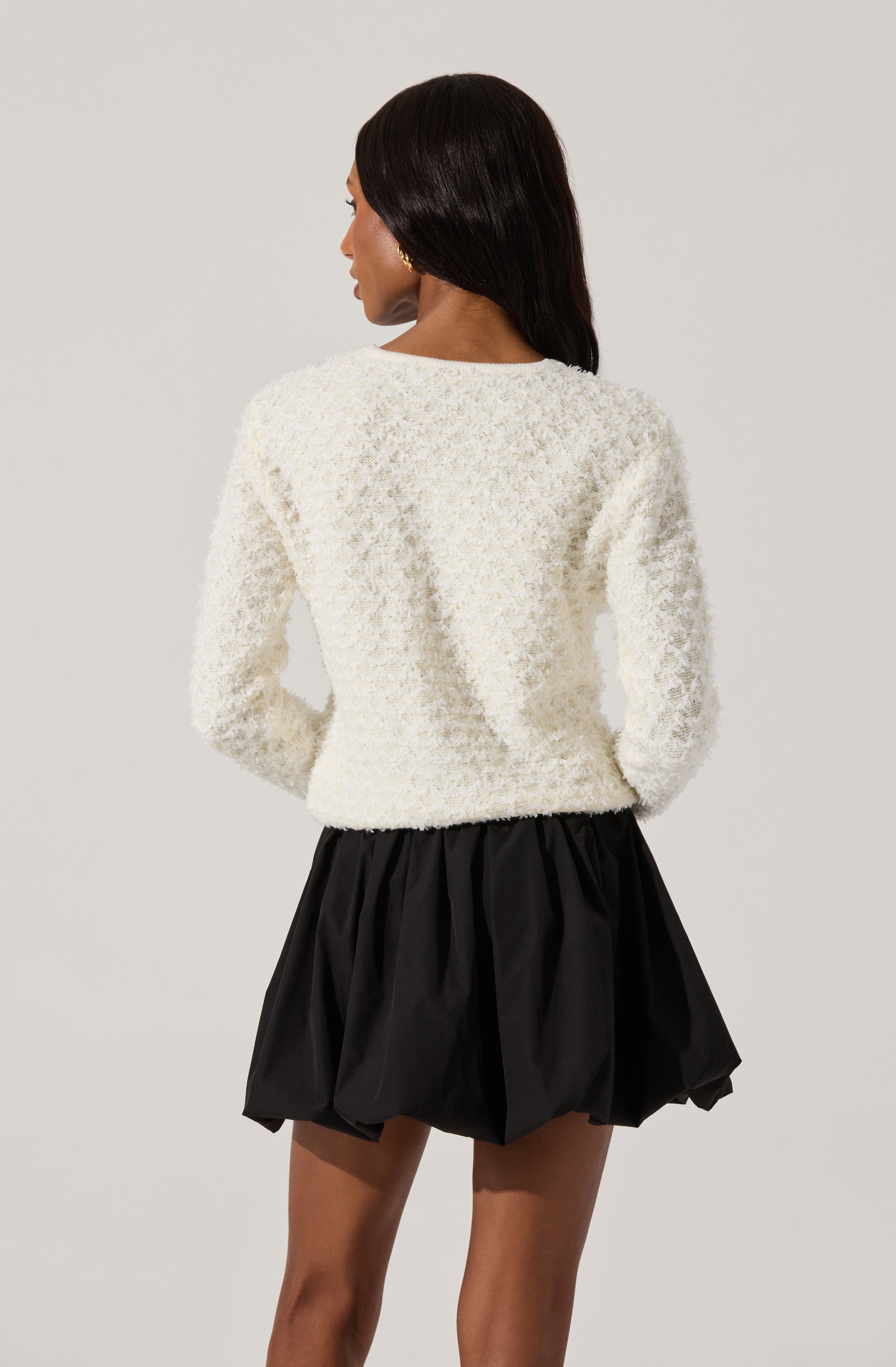 A woman is wearing a textured, long-sleeved, cream-colored sweater and a black, puffed skirt with a gathered waistband.