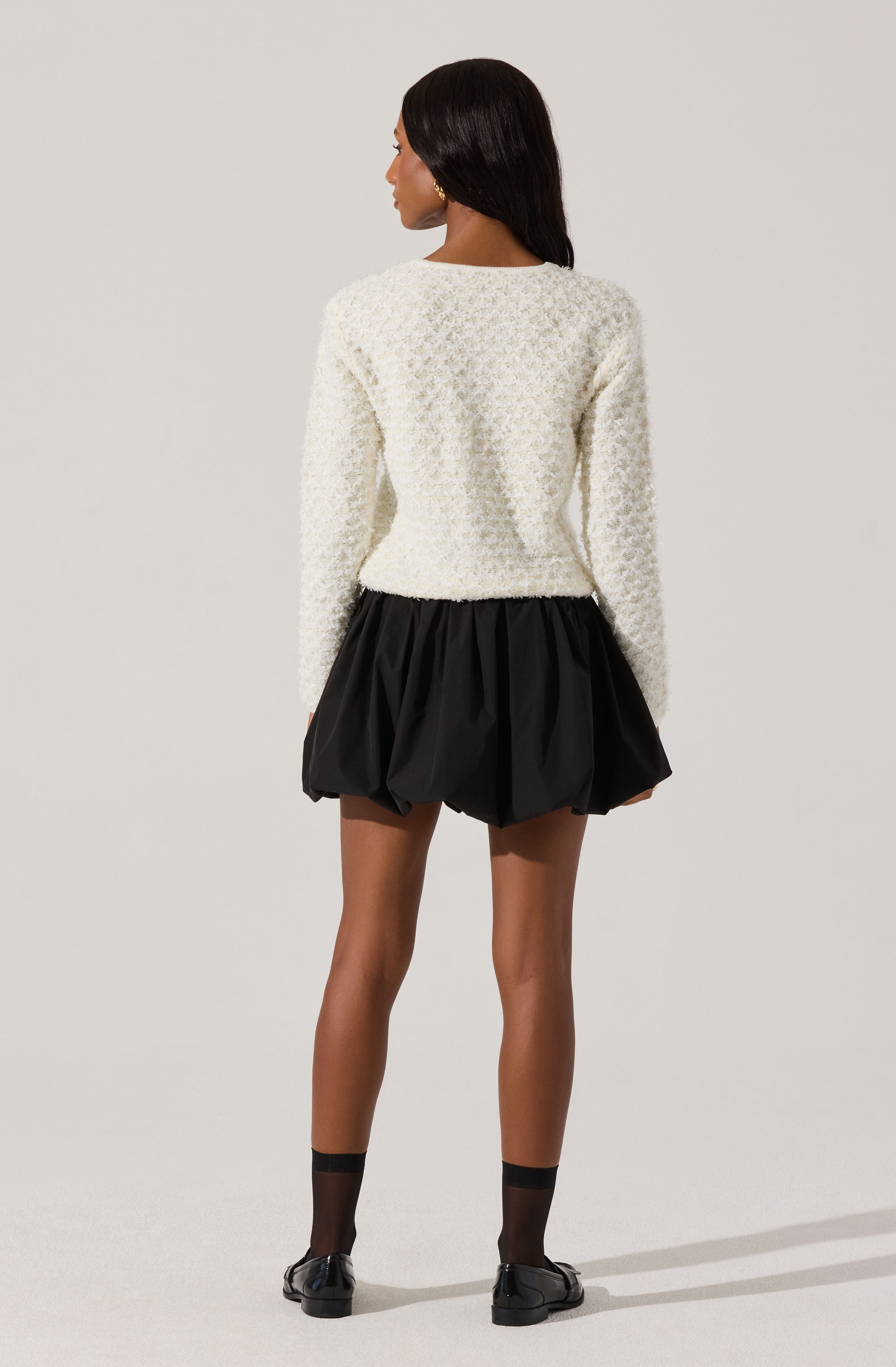 A woman is wearing a white, textured, long-sleeve sweater with a rounded neckline. She pairs it with a black, puffy, balloon-style skirt that extends to mid-thigh. She has on black ankle socks and black shoes. She has dark, straight hair and is standing with her back to the camera against a plain, light-colored background.