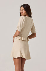 A woman is wearing a light beige outfit consisting of a short-sleeved, pleated top with puffed sleeves and a matching skirt. The top has a fitted bodice with vertical seams and a peplum hem that flares slightly at the waist. The skirt is high-waisted, slightly flared, and above knee-length.