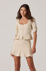 A woman is wearing a cream-colored two-piece outfit consisting of a fitted top with short, puffed sleeves and a peplum hem, featuring vertical seams and a row of buttons down the front. The skirt is short, high-waisted, with a slight A-line shape and subtle pleats at the hem. She is accessorized with gold hoop earrings and a ring on her right hand.