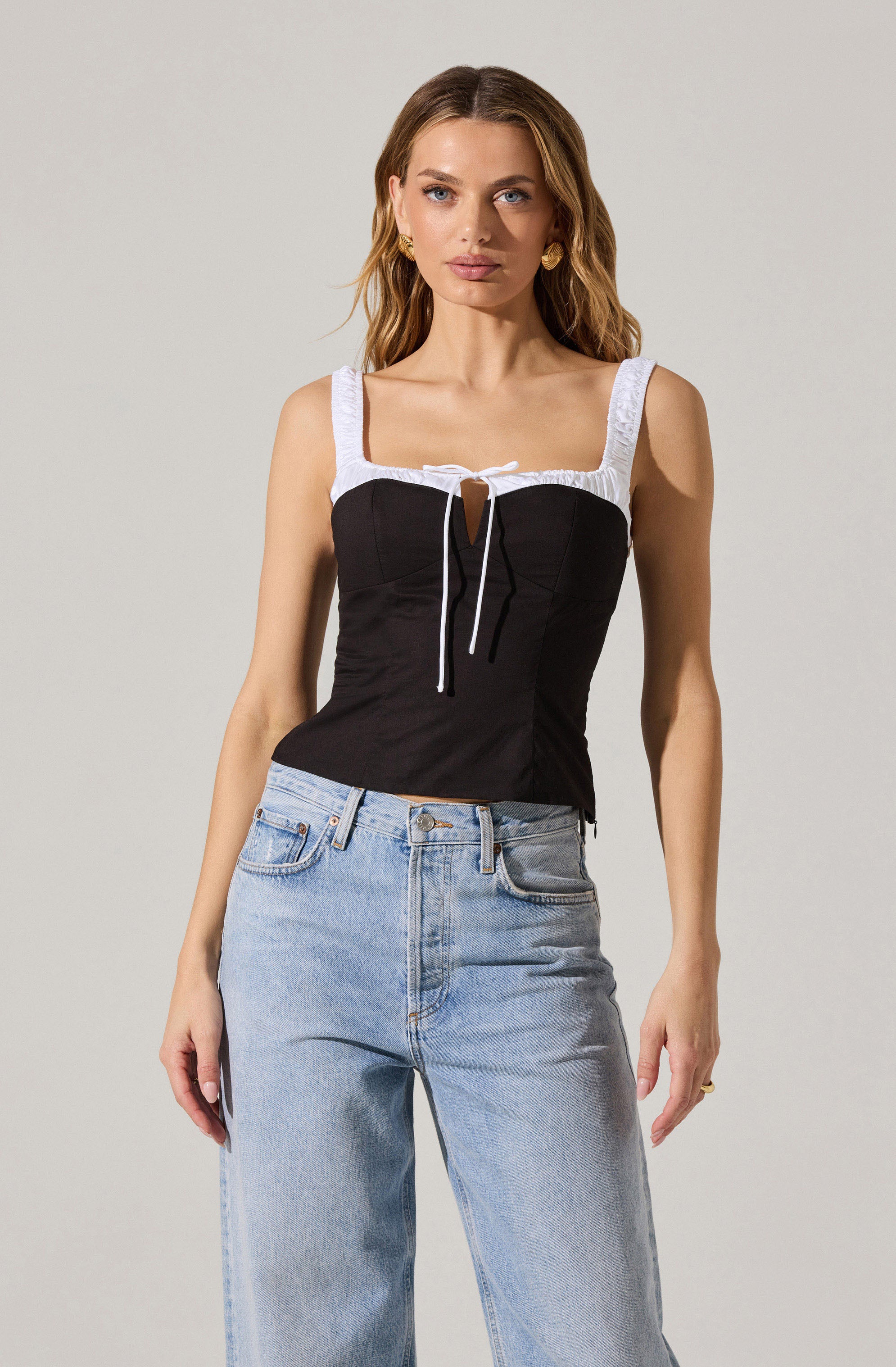 A woman is wearing a sleeveless top with wide white straps and a black bodice featuring a front keyhole cutout tied with a white string. She pairs the top with high-waisted, light-wash jeans.