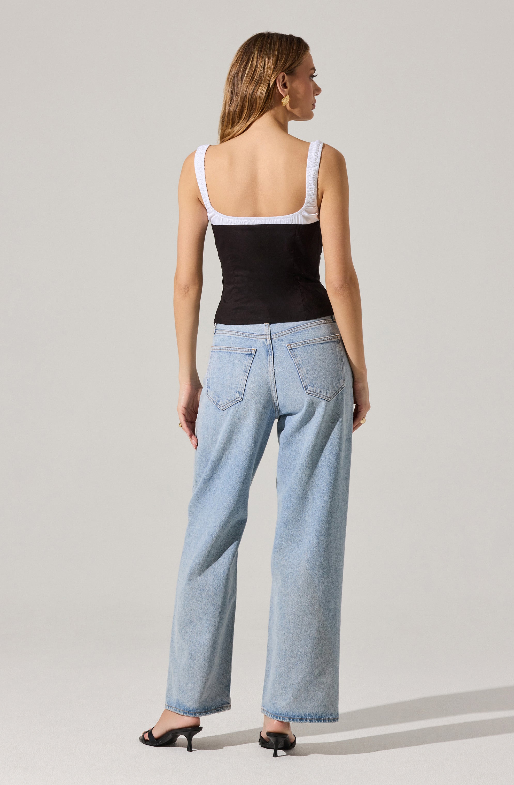 A woman is wearing a sleeveless top with wide straps and a square neckline. The top is black with white straps and trim along the neckline and armholes. She is also wearing light blue, high-waisted, wide-leg jeans with visible stitching and back pockets, and black heeled shoes.