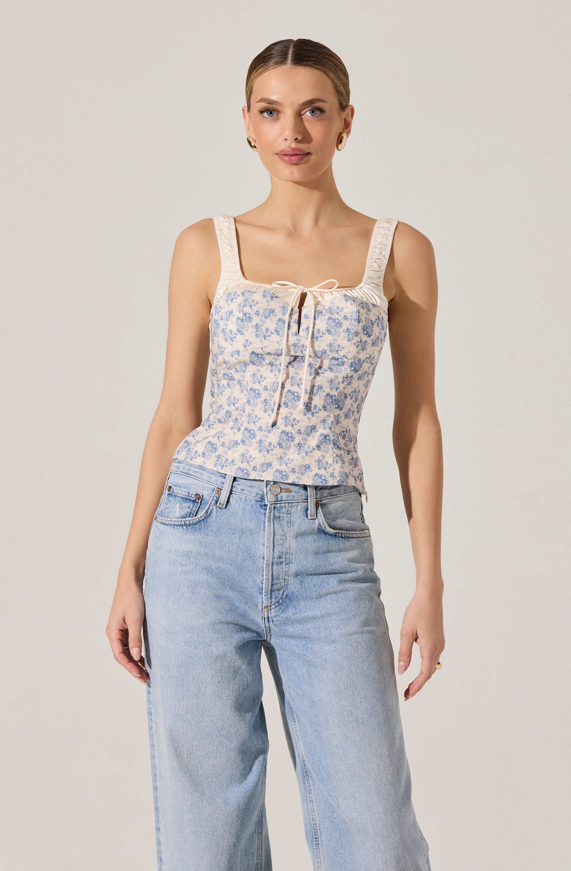 A woman is wearing a sleeveless, fitted top with wide straps. The top features a delicate floral pattern in blue and beige, with a cream-colored bow at the neckline and a tie detail in the center. She pairs it with high-waisted, light-wash, straight-leg jeans.