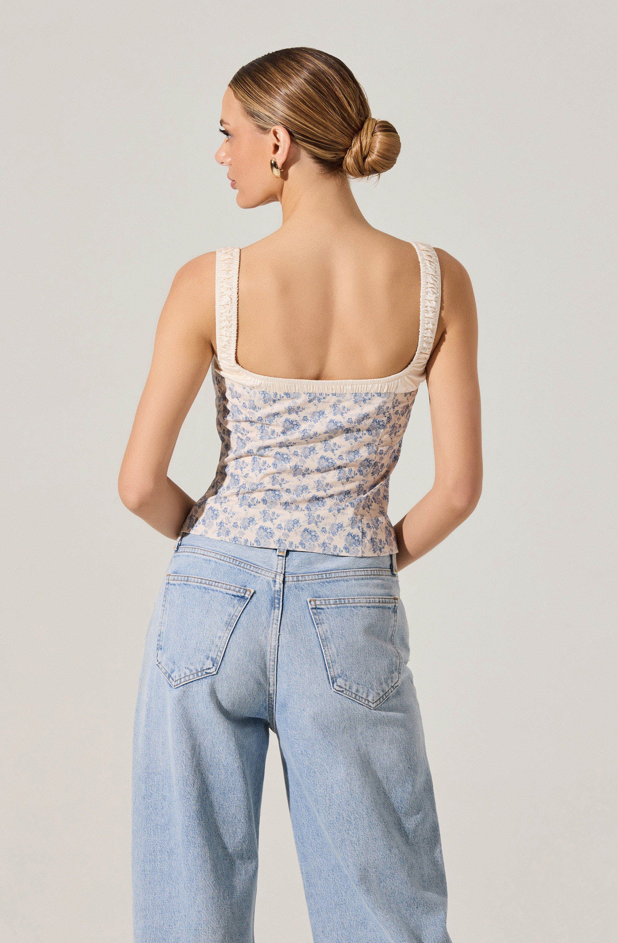 A woman is wearing a sleeveless top with wide straps, featuring a floral pattern in shades of blue and cream, with lace trim along the edges of the straps and neckline. She is also wearing high-waisted, light-wash jeans with a relaxed fit and visible back pockets. Her hair is styled in a low, neat bun.