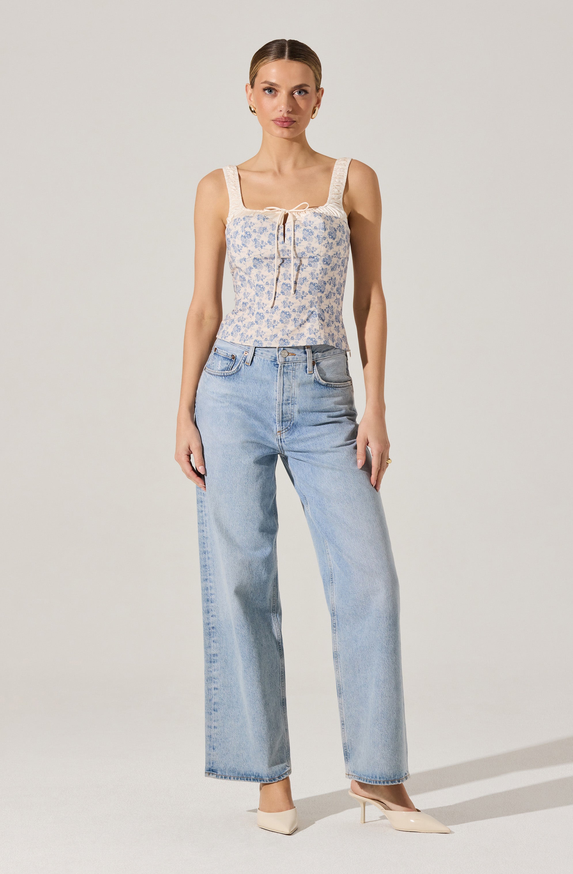 A woman is wearing a sleeveless, cream-colored top with a light blue floral pattern and a tie detail at the neckline. She pairs this with high-waisted, light-wash, wide-leg jeans and beige pointed-toe heels.