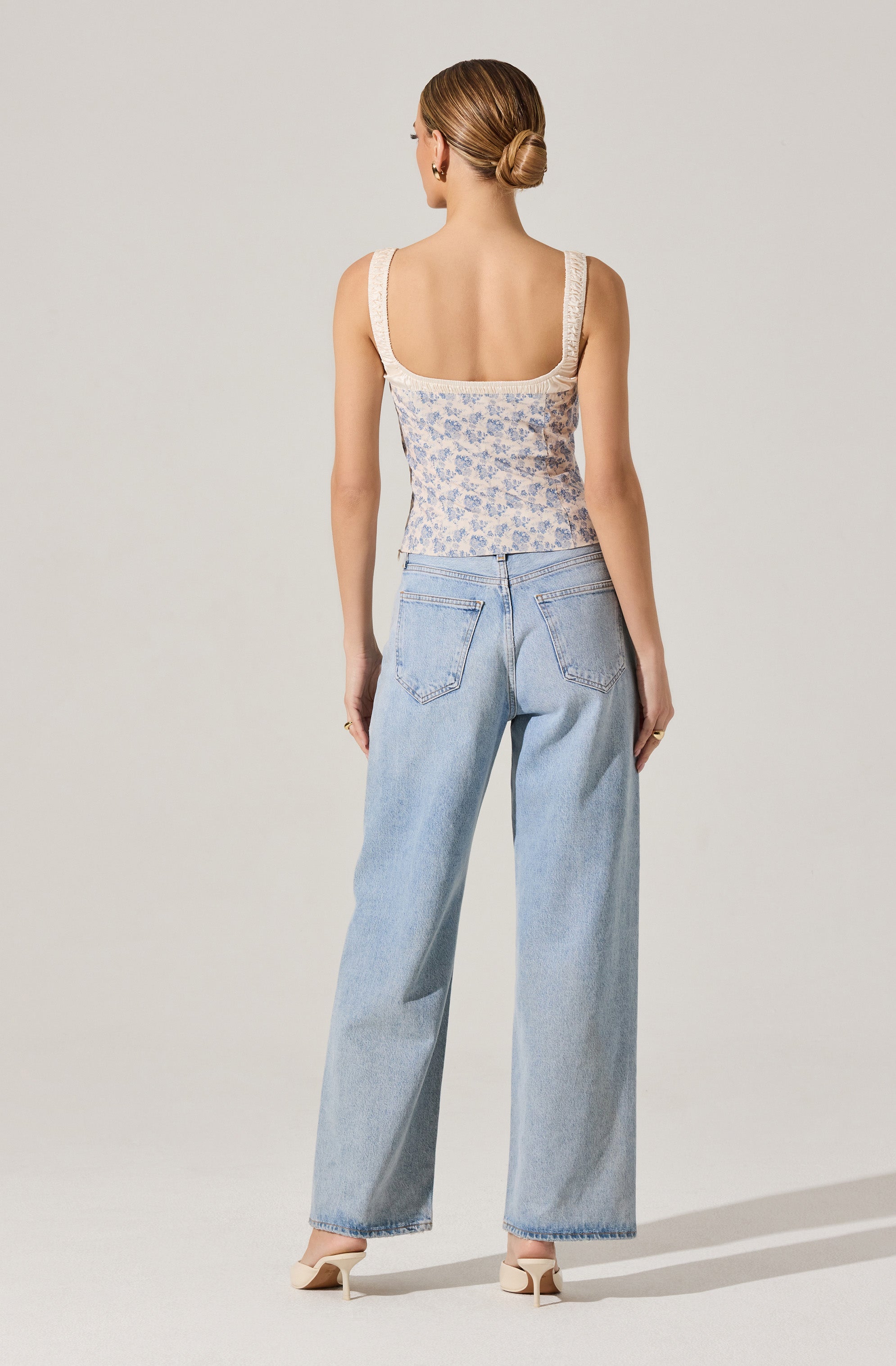 A woman is wearing a sleeveless, fitted top with wide straps, featuring a light-colored floral pattern and lace details on the straps. She is also wearing high-waisted, light-wash wide-leg jeans with back pockets, and beige heeled shoes. Her hair is styled in a low bun.