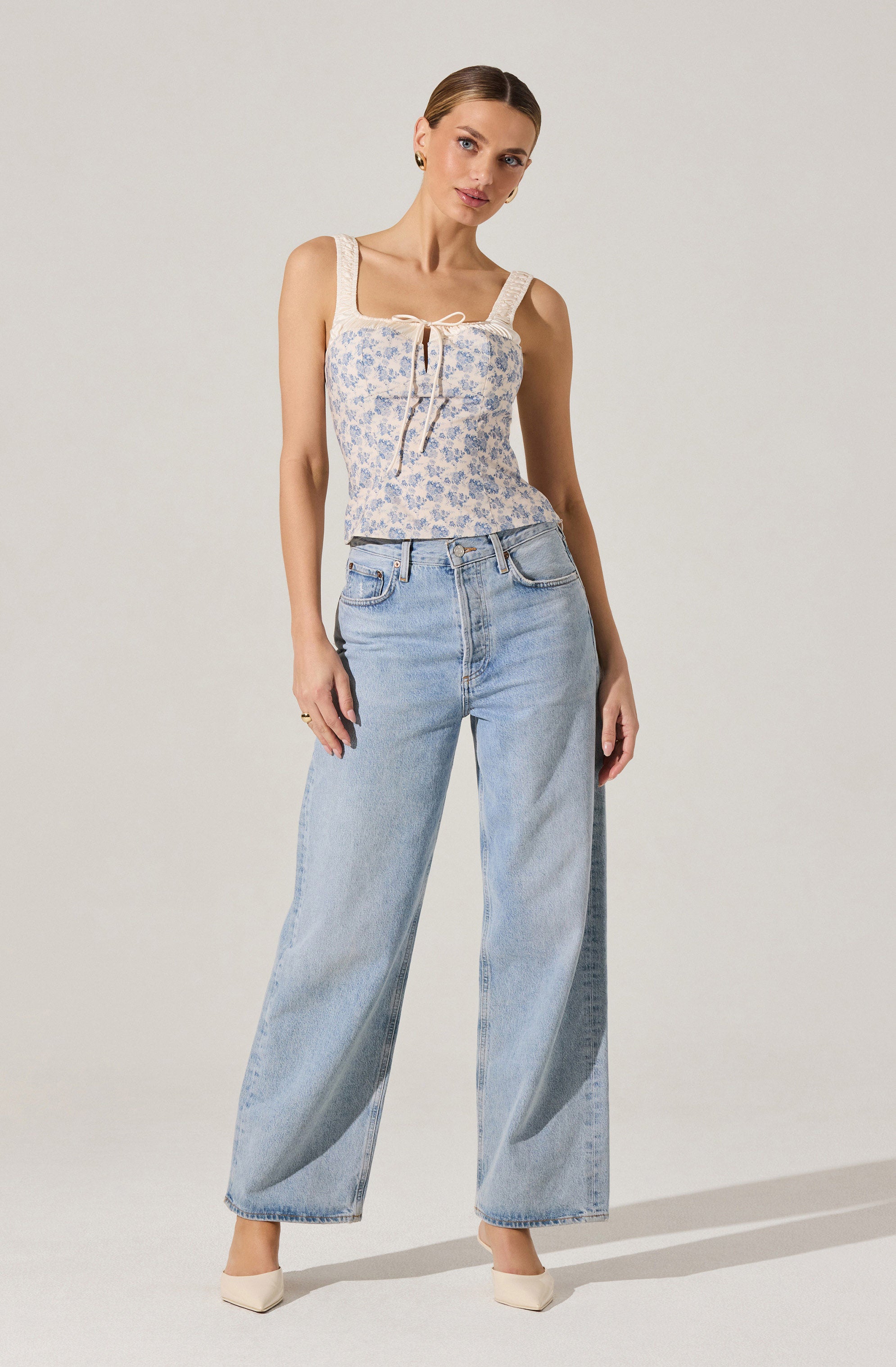 A woman is wearing a sleeveless top with wide shoulder straps, featuring a delicate floral pattern in light colors and a tied bow detail at the front. She is also wearing high-waisted, wide-leg light blue jeans. The outfit is completed with light-colored pointed-toe shoes.