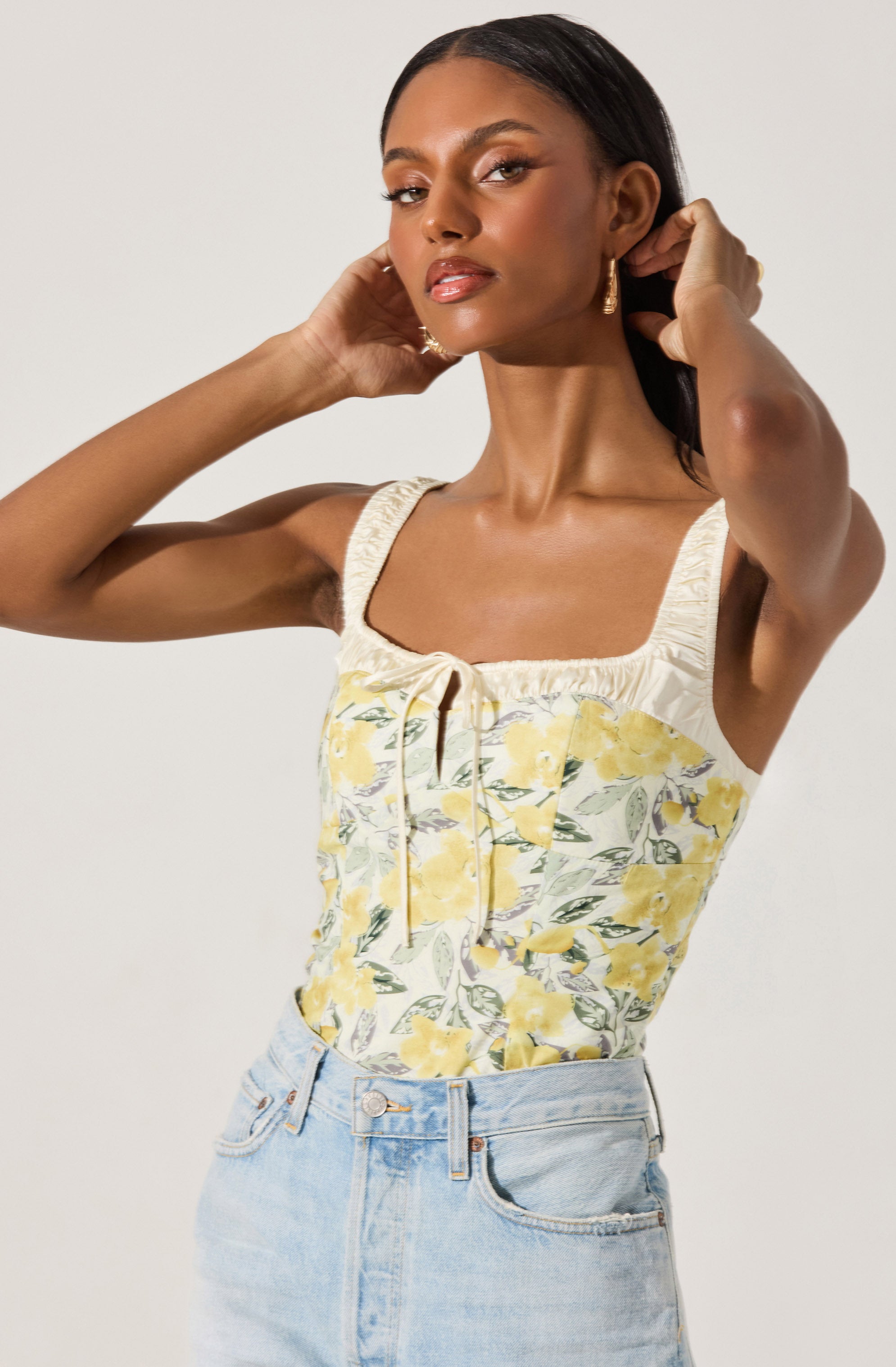 A woman is wearing a sleeveless, square-neck top with a floral pattern featuring yellow flowers, green leaves, and grey accents. The top has a gathered neckline with a small bow tie at the center. She pairs it with light-wash denim jeans that sit at her waist.