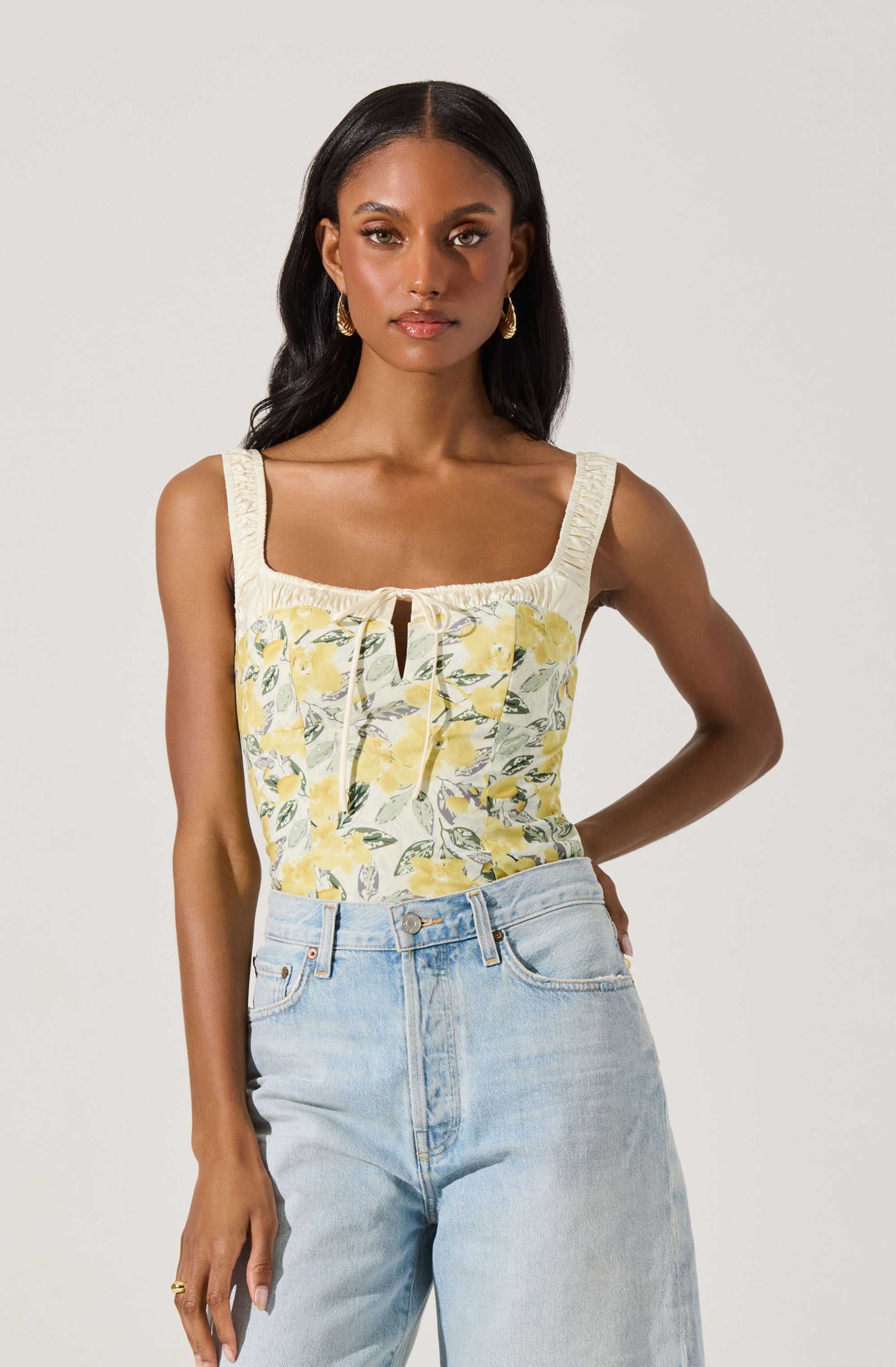 A woman is wearing a sleeveless top with wide straps and a square neckline, featuring a floral pattern with yellow flowers and green leaves on a light background. The top has a small keyhole opening at the center front, with a narrow tie. The woman pairs this with high-waisted, light-wash denim jeans.