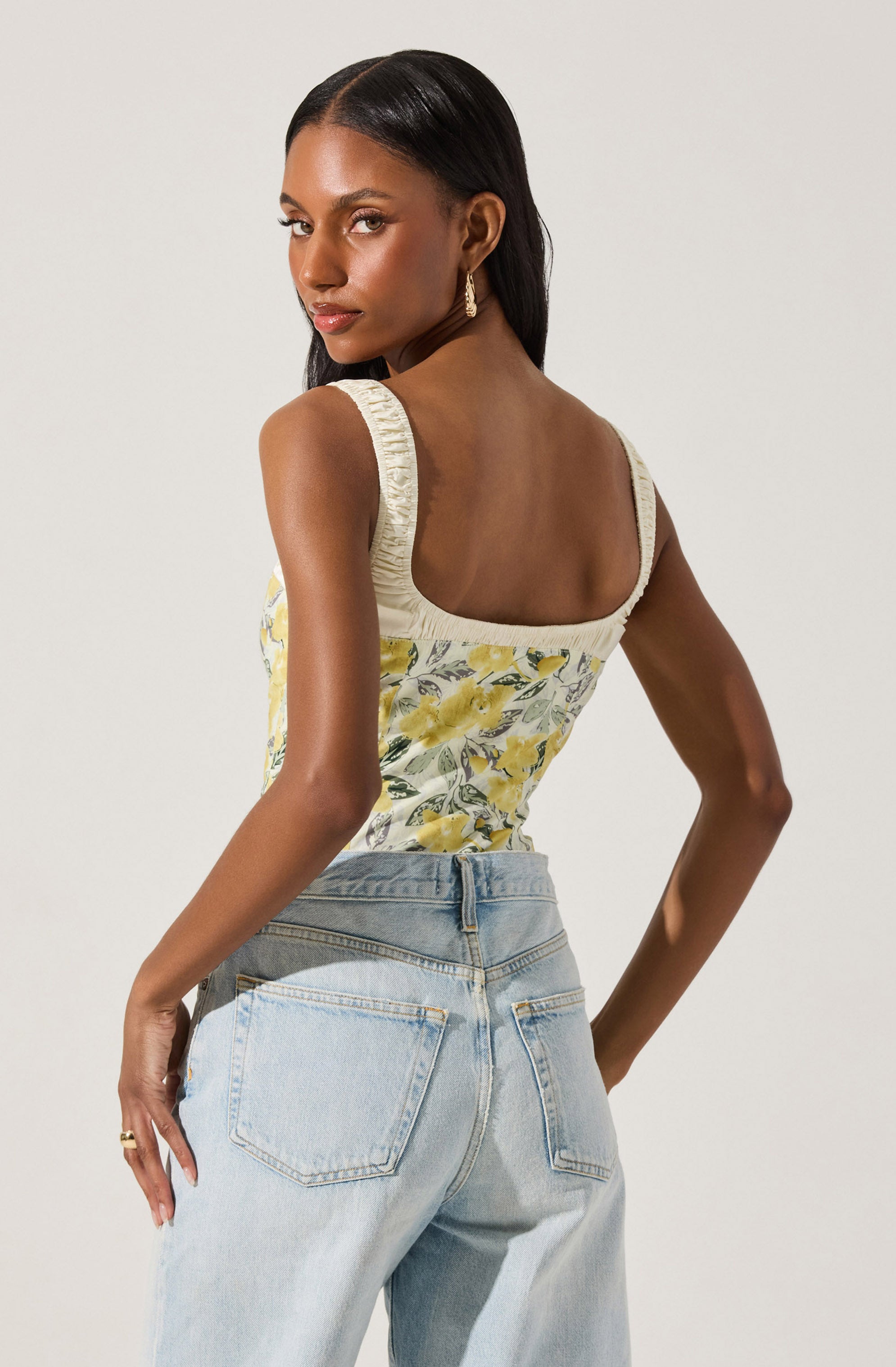 A woman is wearing a sleeveless, fitted top with a floral print in shades of yellow, white, and gray, featuring ruching detail on the straps. She pairs it with high-waisted, light-wash jeans that have a relaxed fit and classic five-pocket styling.