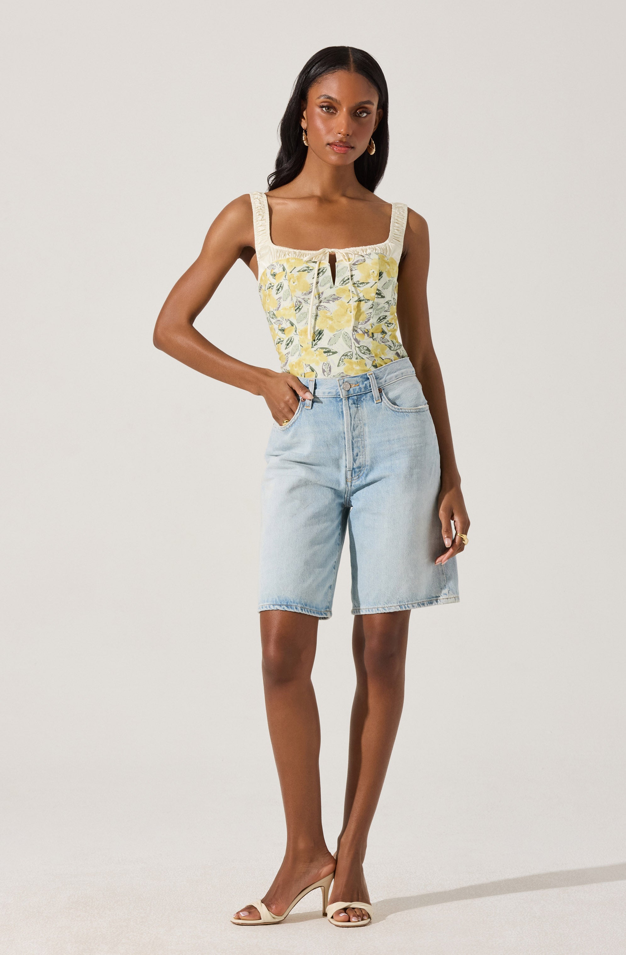 A woman is wearing a sleeveless, square-neck top with a light yellow background and a floral pattern in shades of yellow and green, with lace trim along the top edge. She is also wearing light wash denim knee-length shorts and beige high-heeled sandals.