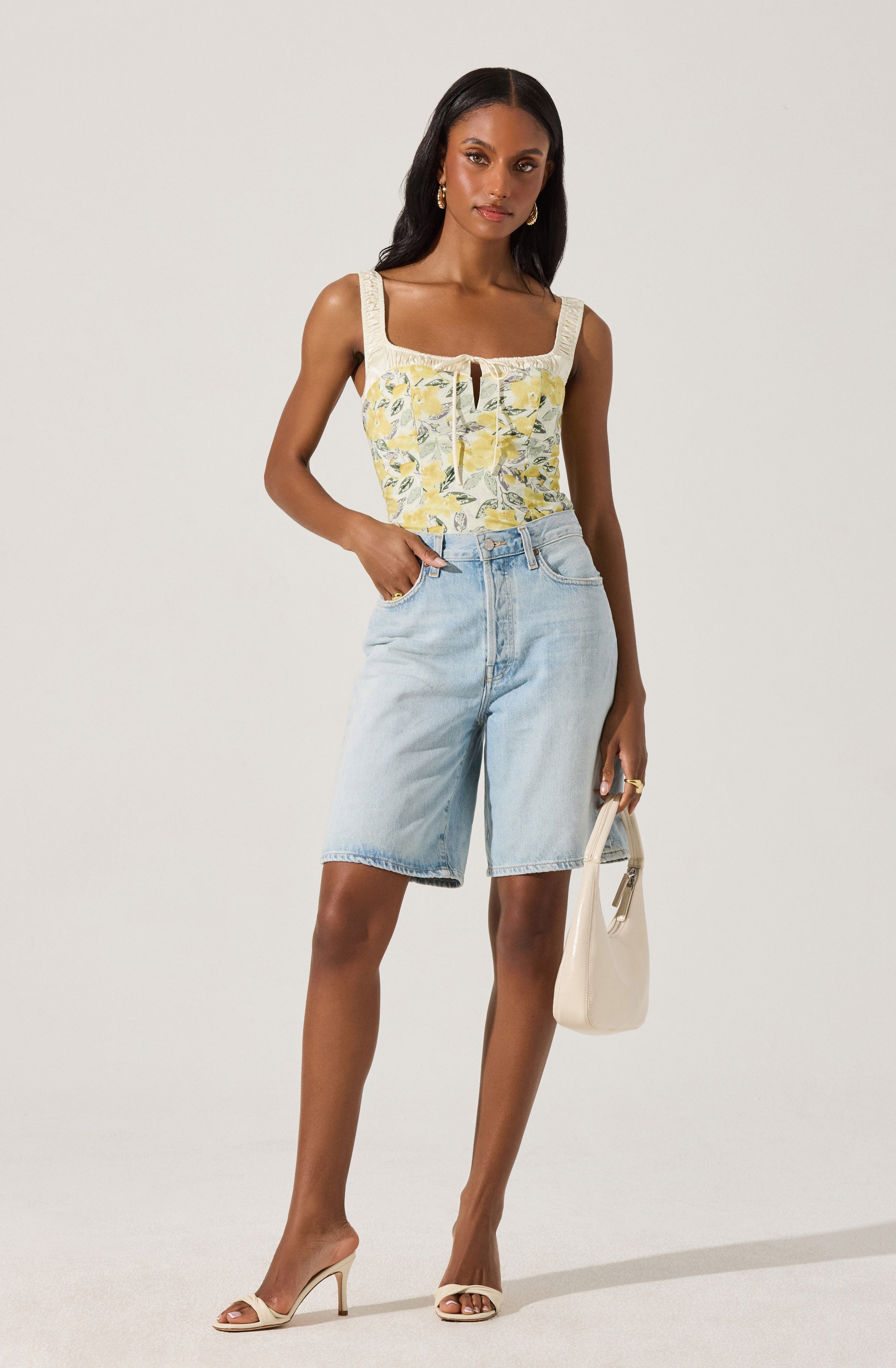 A woman is wearing a sleeveless, square-neck top with a yellow floral pattern and beige lace trim on the straps. She pairs the top with light blue high-waisted denim knee-length shorts. She accessorizes with gold hoop earrings, multiple rings, and carries a white handbag. She is also wearing beige open-toe high heels.
