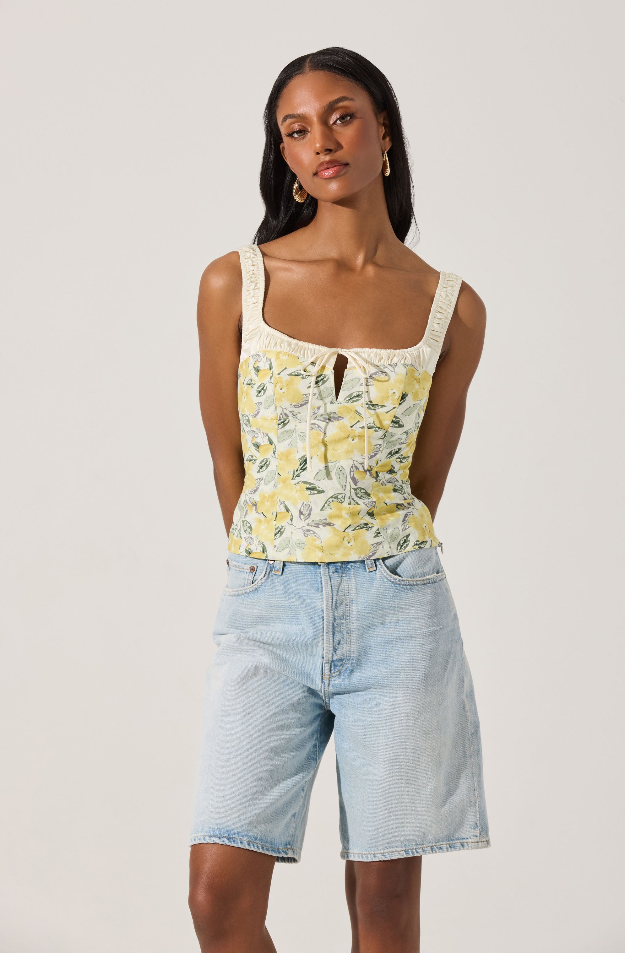 A woman is wearing a sleeveless top with wide straps, a square neckline, and a keyhole detail at the center front. The top features a floral pattern with yellow and green leaves on a light background. She pairs it with light-wash denim shorts.