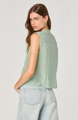 A woman is wearing a sleeveless, light green top with embroidered and cut-out details along the seams and yoke. The top has a relaxed fit and a ruffled neckline. She pairs it with high-waisted, light-wash denim jeans. She has long, wavy brown hair and is accessorized with small hoop earrings.