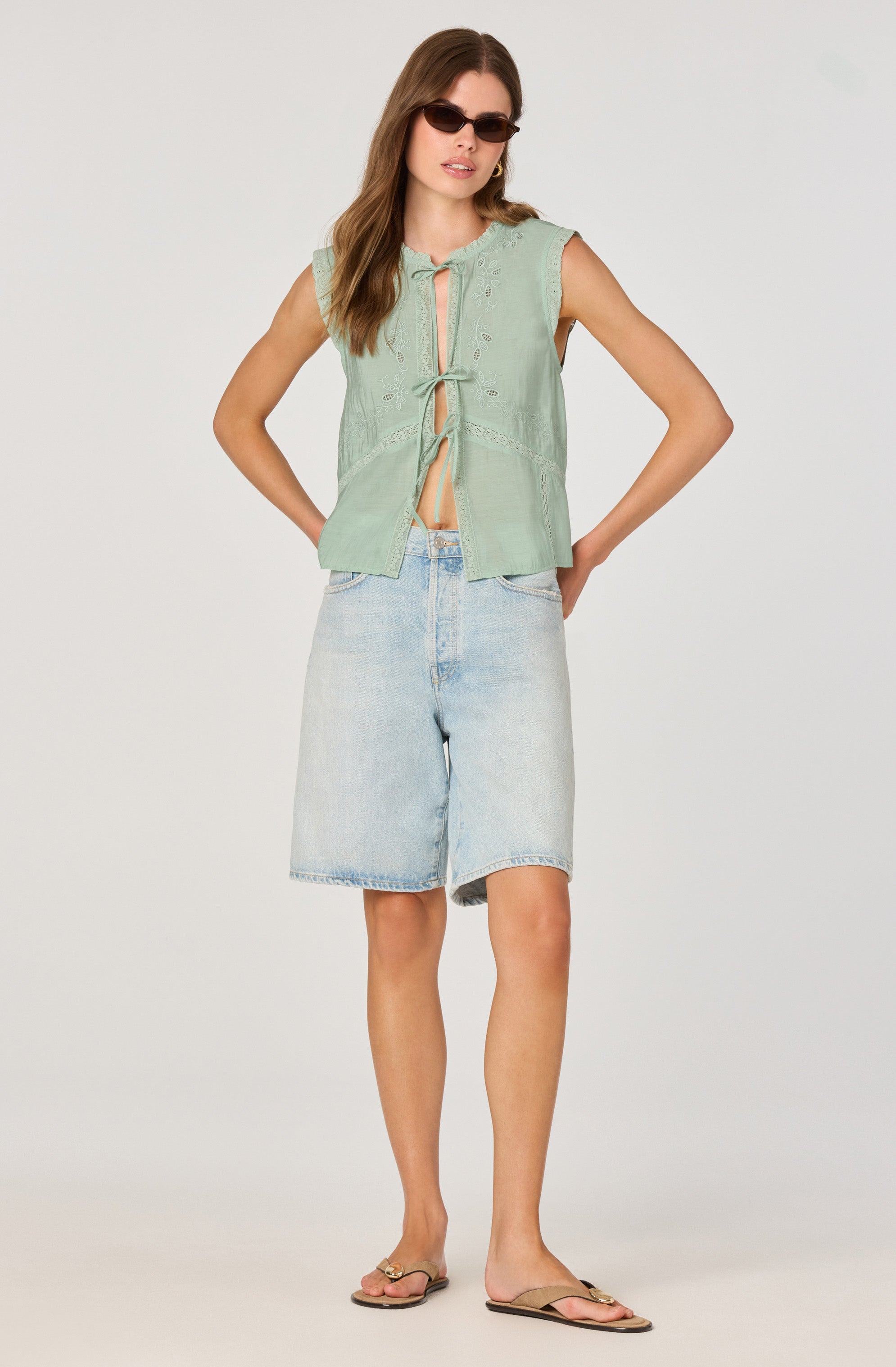 A woman is wearing a sleeveless, light green embroidered top with a front opening tied with a bow, paired with light-wash denim shorts that reach just above the knees. She accessorizes with dark sunglasses and gold hoop earrings, and she is wearing flat sandals.