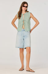A woman is wearing a sleeveless, light green embroidered top with a front opening tied with a bow, paired with light-wash denim shorts that reach just above the knees. She accessorizes with dark sunglasses and gold hoop earrings, and she is wearing flat sandals.