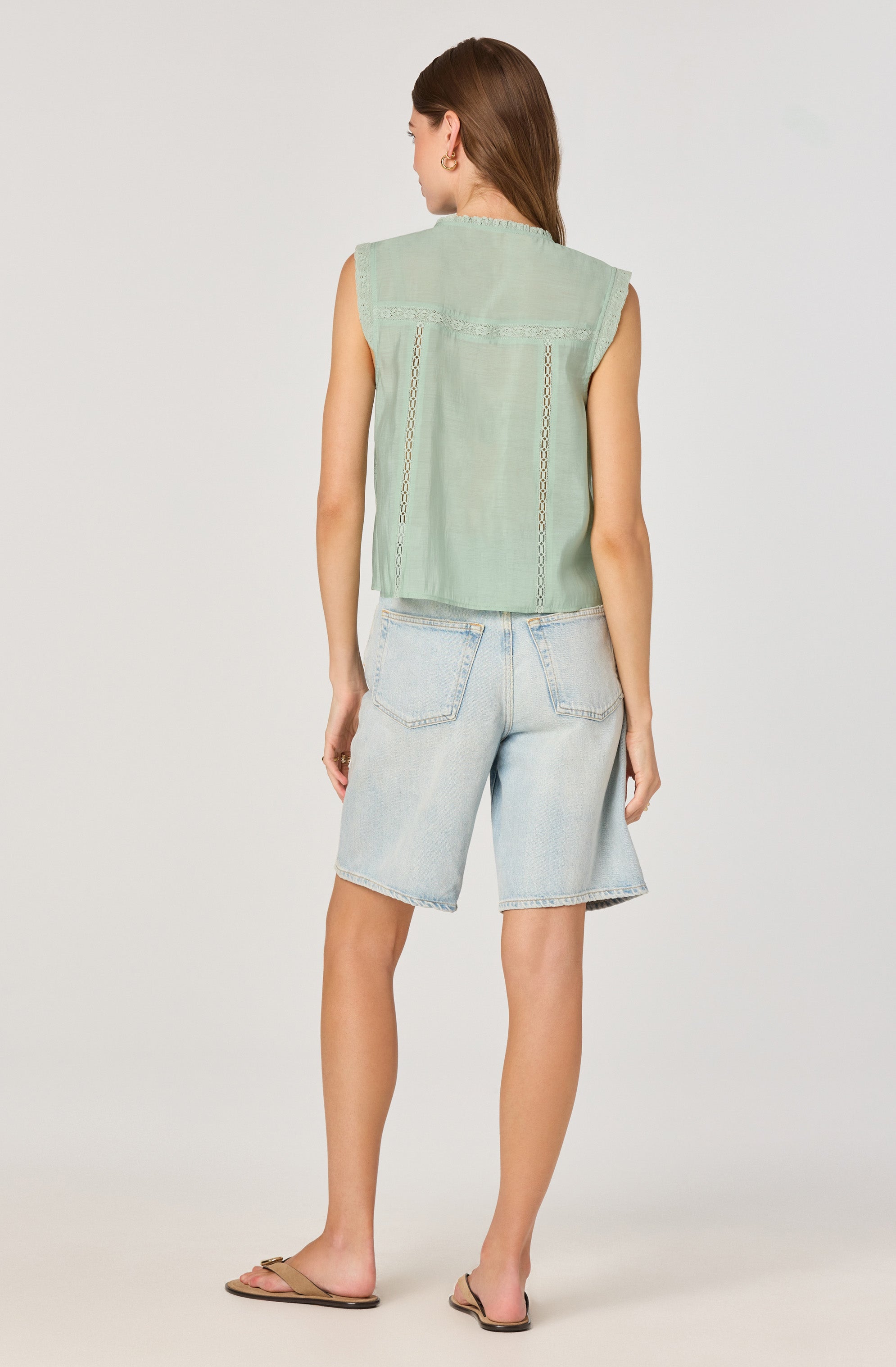A woman is wearing a sleeveless, light green top with decorative stitching and lace details. She pairs it with light-wash denim shorts that are knee-length and have large back pockets. She is also wearing tan flat sandals.