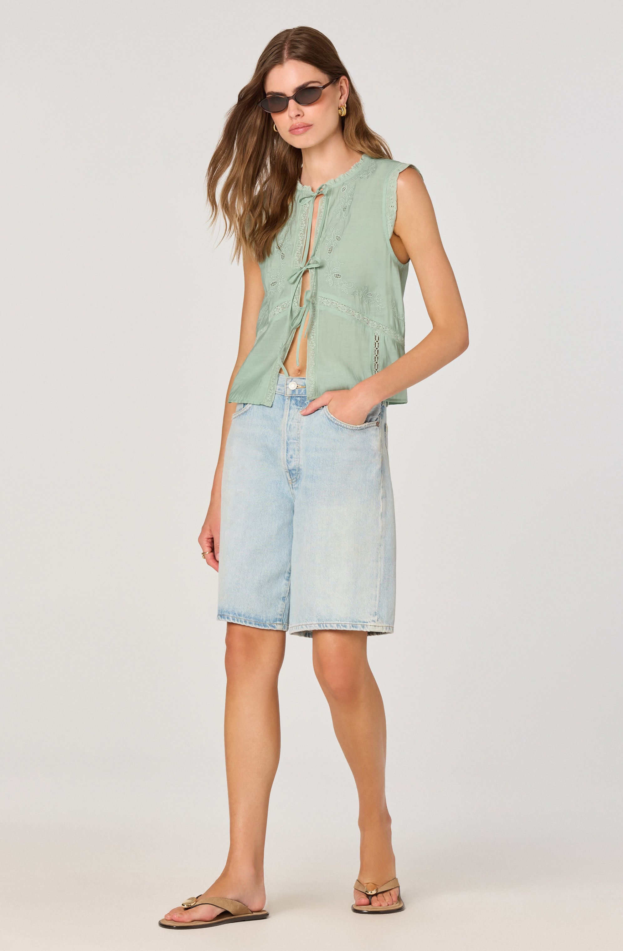 A woman is wearing a sleeveless, light green, button-up top with a tie detail at the front and embroidered accents. She pairs it with light-washed, knee-length denim shorts. She accessorizes with dark sunglasses and gold earrings, and she is wearing tan sandals.