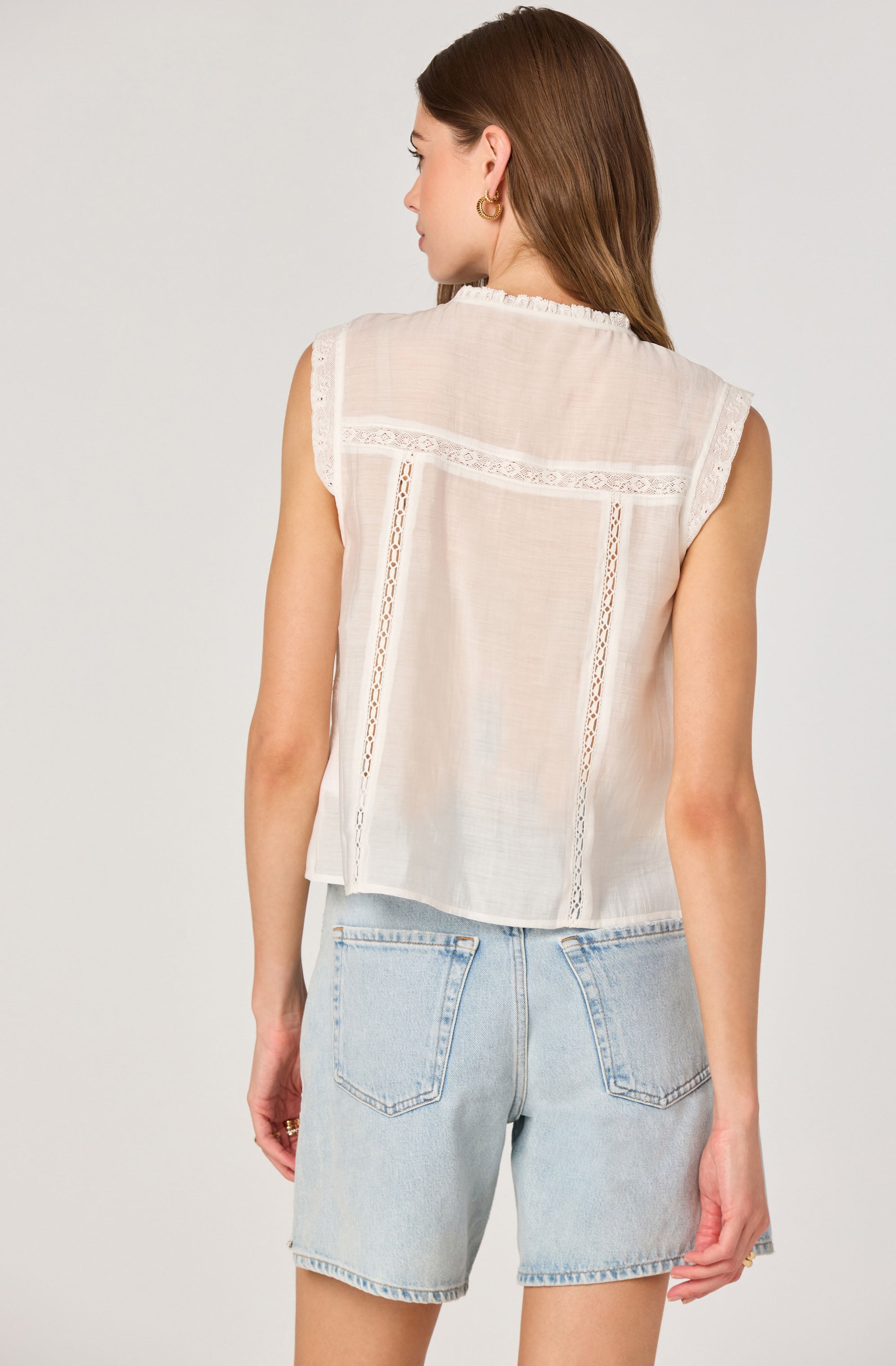 A woman is wearing a sleeveless, light-colored, semi-sheer blouse with lace and crochet details along the back and shoulders. The blouse has a relaxed fit with a frilled neckline. She is also wearing light blue denim shorts with a high waist and back pockets.