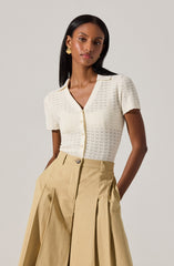 A woman is wearing a short-sleeved, cream-colored knitted top with a subtle V-neckline and buttons down the front. She pairs it with high-waisted, beige wide-leg pants that feature pleats and large front pockets. She accessorizes with gold hoop earrings.
