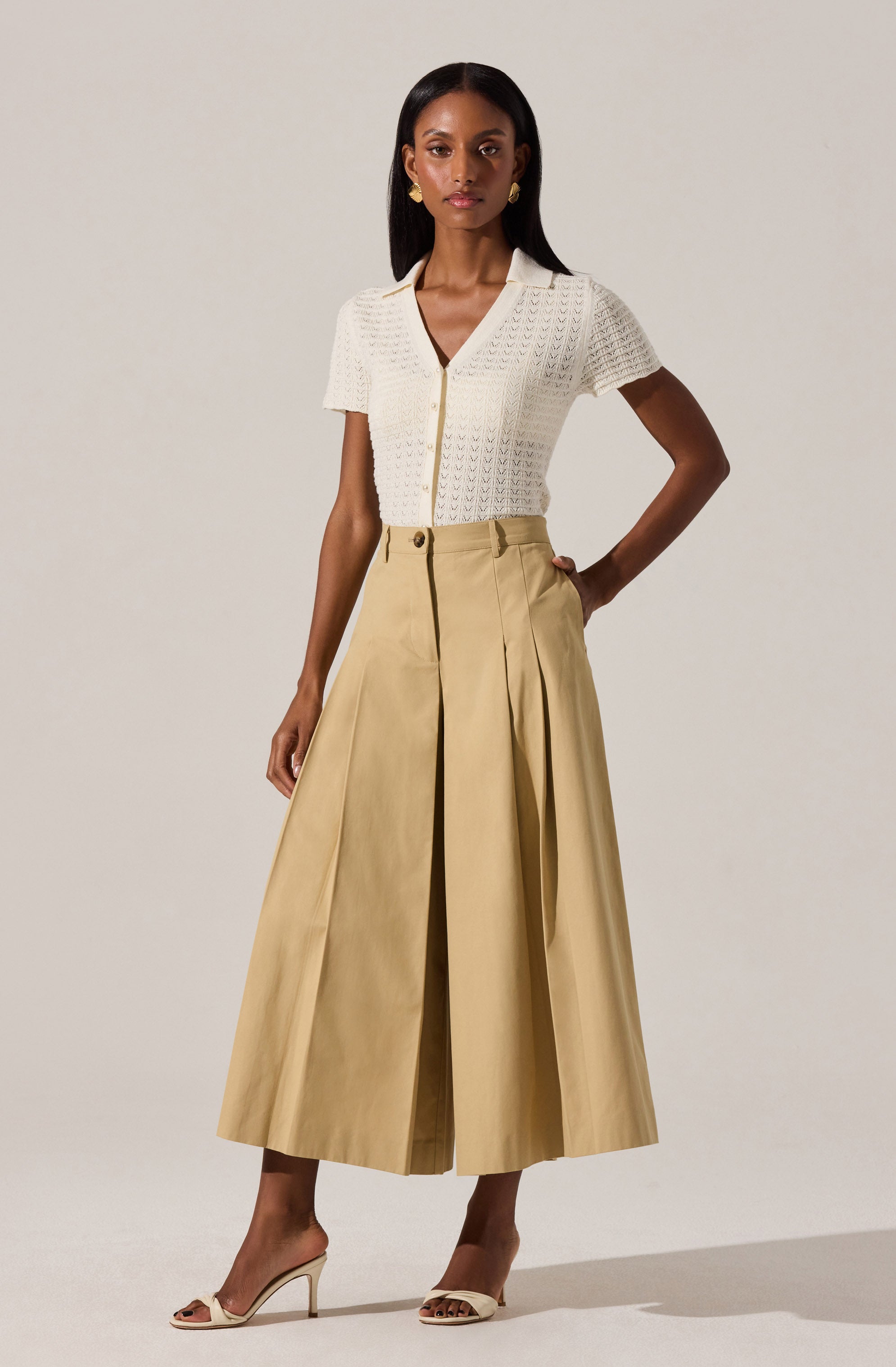 A woman is wearing a short-sleeved, textured, cream-colored knit top with a V-neckline and buttons down the front. She pairs it with high-waisted, wide-leg, light beige trousers that feature pleats and a front zipper.