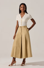 A woman is wearing a short-sleeved, textured, cream-colored knit top with a V-neckline and buttons down the front. She pairs it with high-waisted, wide-leg, light beige trousers that feature pleats and a front zipper.