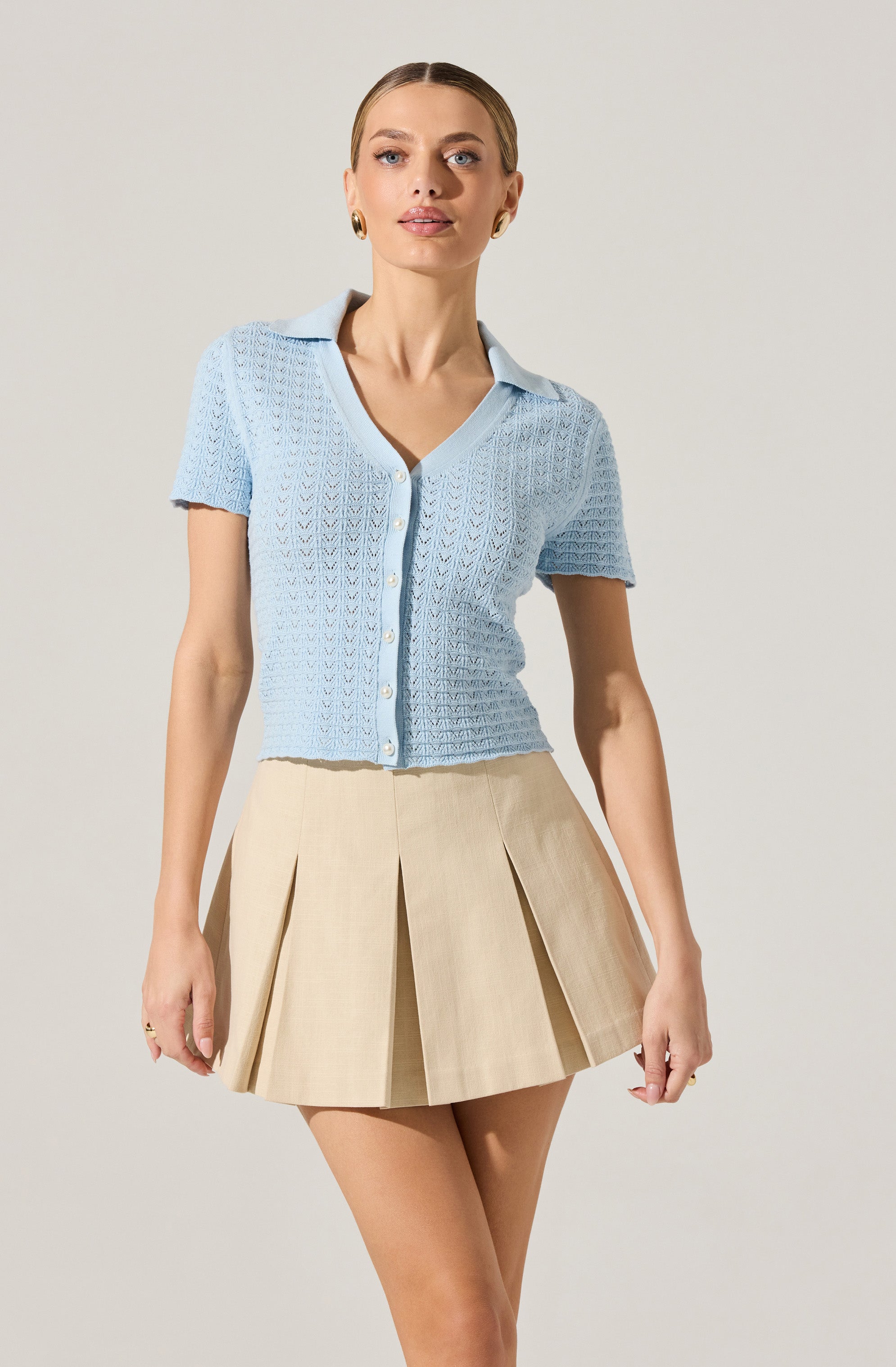 A woman is wearing a light blue, short-sleeved, button-up knit top with a collar. She pairs it with a beige high-waisted pleated skirt.