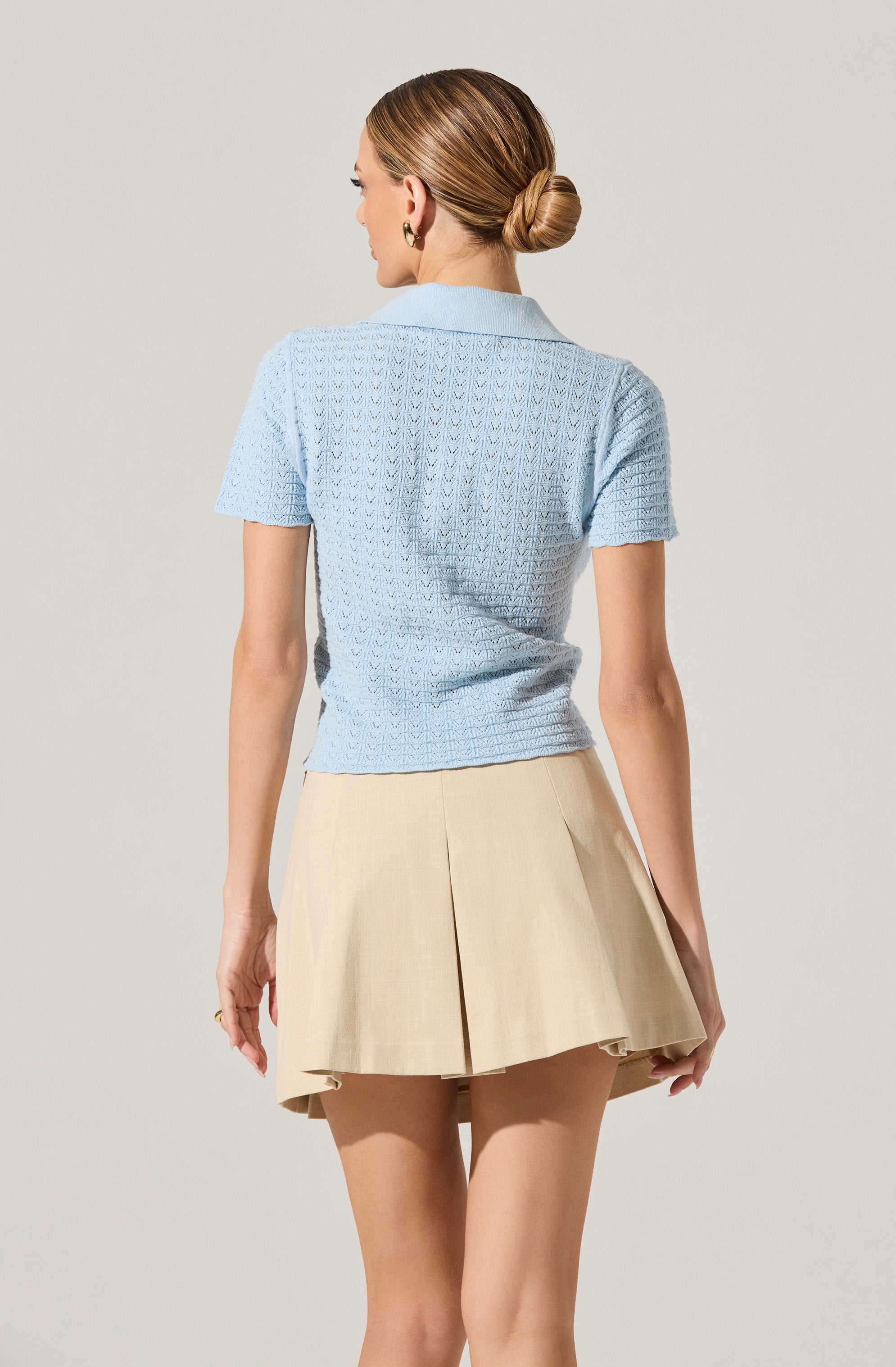 A woman is wearing a light blue short-sleeved knit top with a textured pattern and a light-colored pleated skirt. The top has a classic collar, and the skirt features a front pleat design.