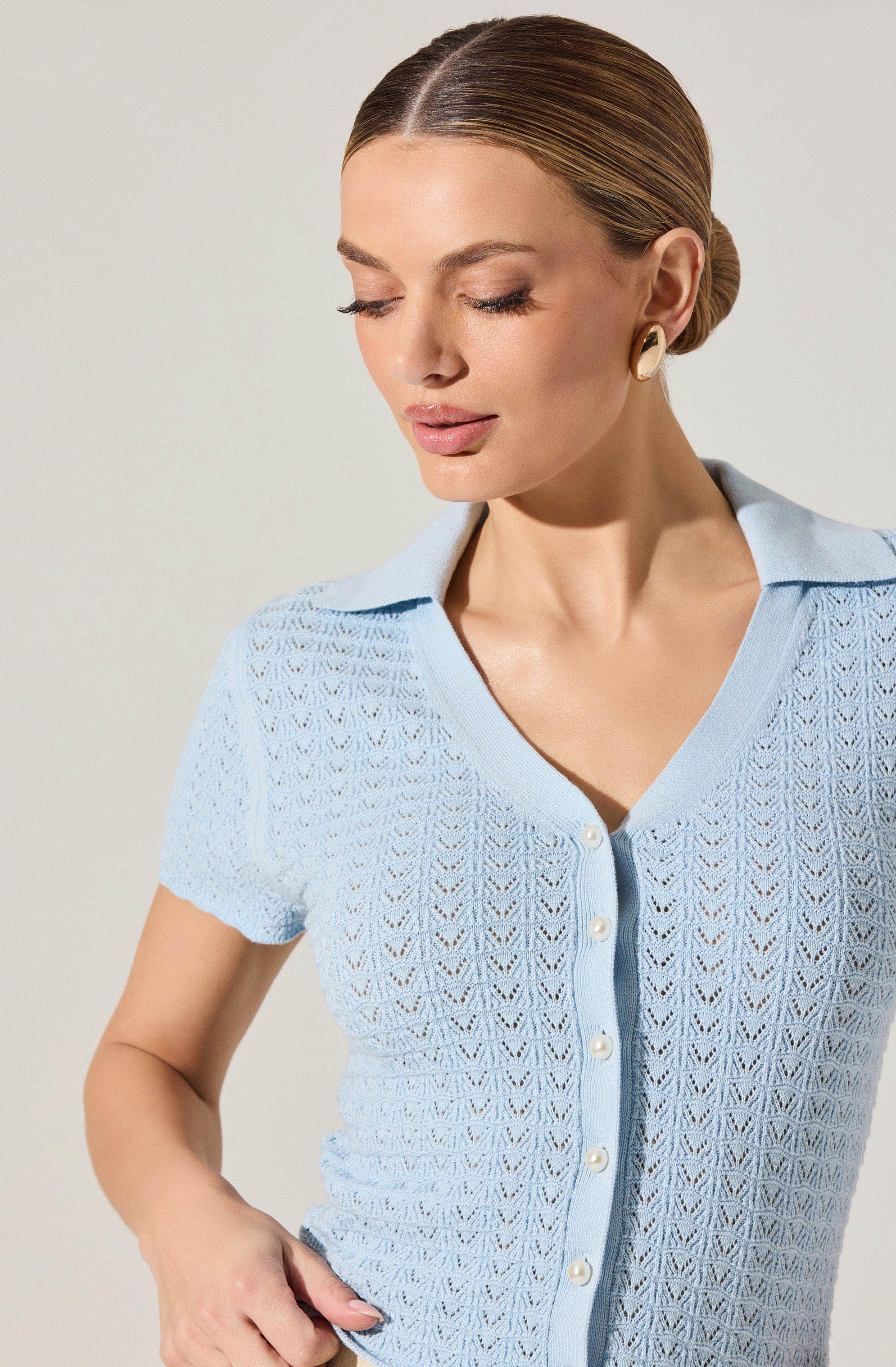 A woman is wearing a light blue, short-sleeved, button-up cardigan with a V-neckline and a collar. The cardigan features a crocheted or lace-like pattern throughout and white buttons down the front.