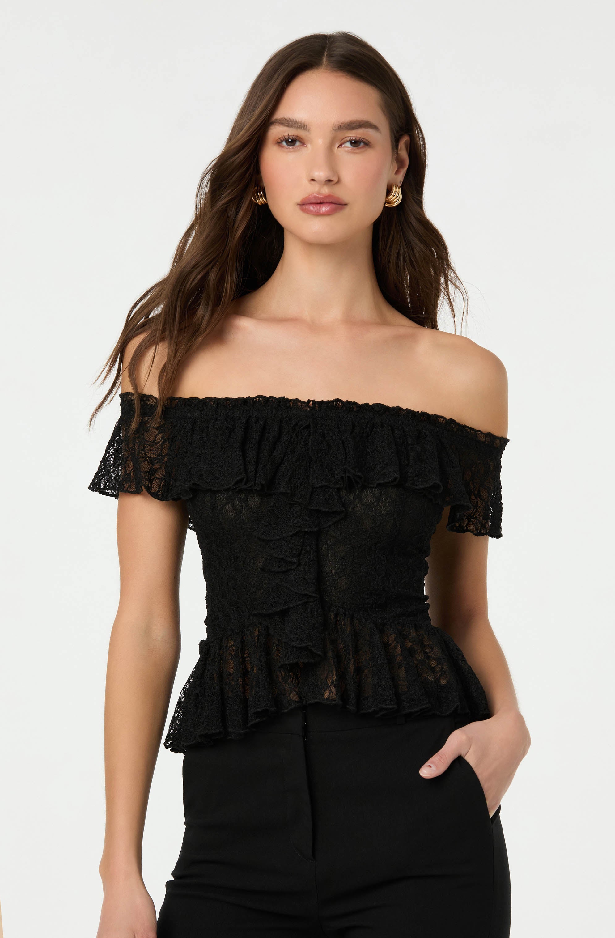 A woman is wearing an off-the-shoulder black lace top with ruffled details around the neckline, sleeves, and hem. The top has a fitted silhouette and features ruffles down the front. She pairs it with black pants and accessorizes with gold hoop earrings.