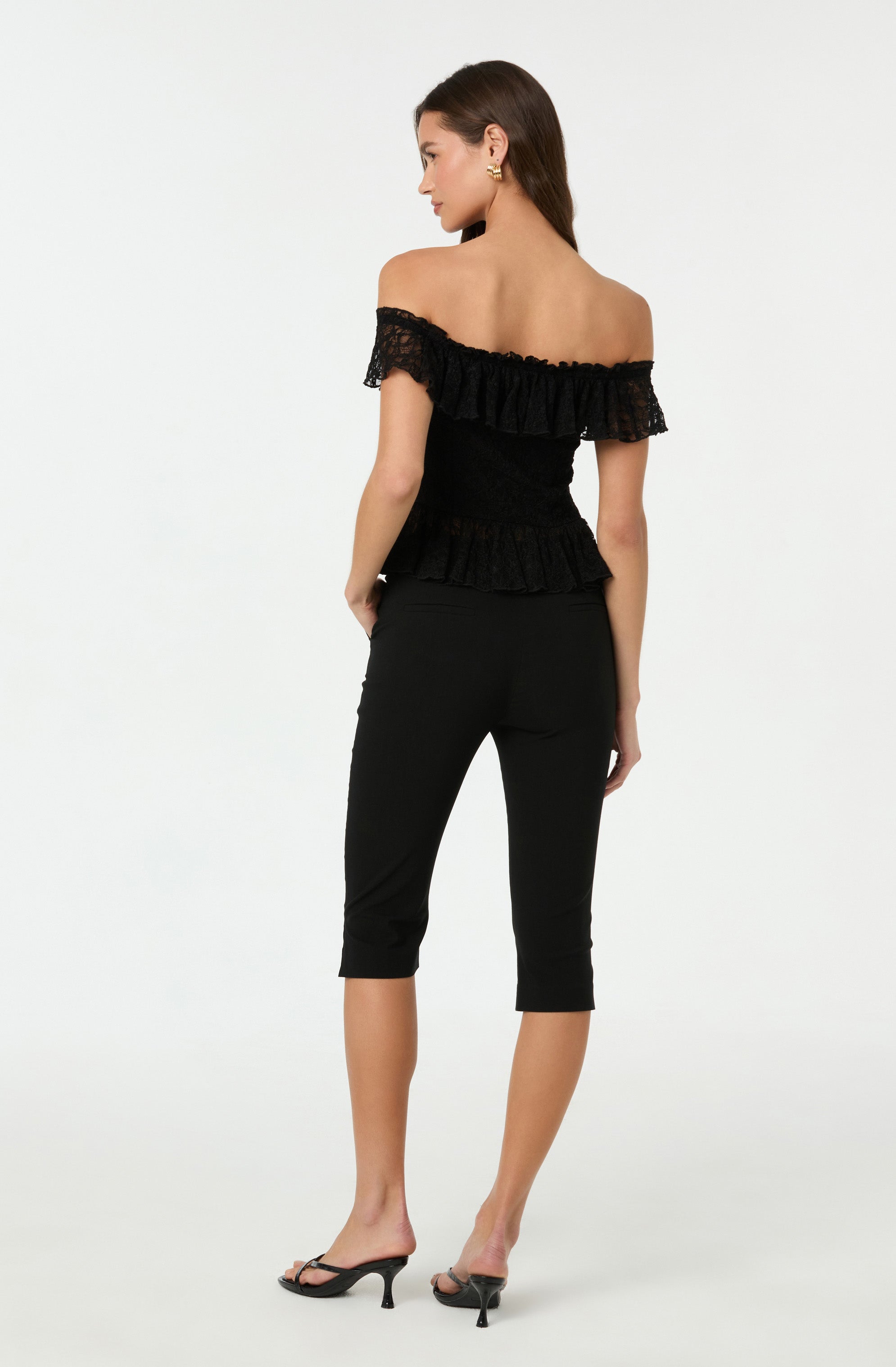 A woman is wearing an off-the-shoulder black top with ruffled lace detailing along the neckline and hem. She pairs it with fitted black cropped pants and black high-heeled sandals.