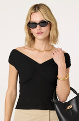 A woman is wearing a black, short-sleeved top with gathered fabric at the shoulders and bust area. She has on black sunglasses, gold hoop earrings, a gold chain necklace, and a gold bracelet. She is holding a black handbag with a gold clasp.
