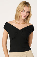 A woman is wearing an off-the-shoulder black top with gathered fabric details at the shoulders and bust. She accessorizes with a gold chain necklace and beige high-waisted pants.