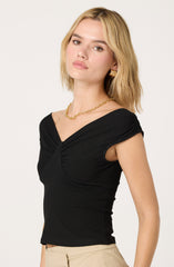 A woman is wearing a fitted black top with gathered detailing at the shoulders and a sweetheart neckline. She pairs it with beige pants and accessorizes with gold jewelry, including earrings and a chain necklace.