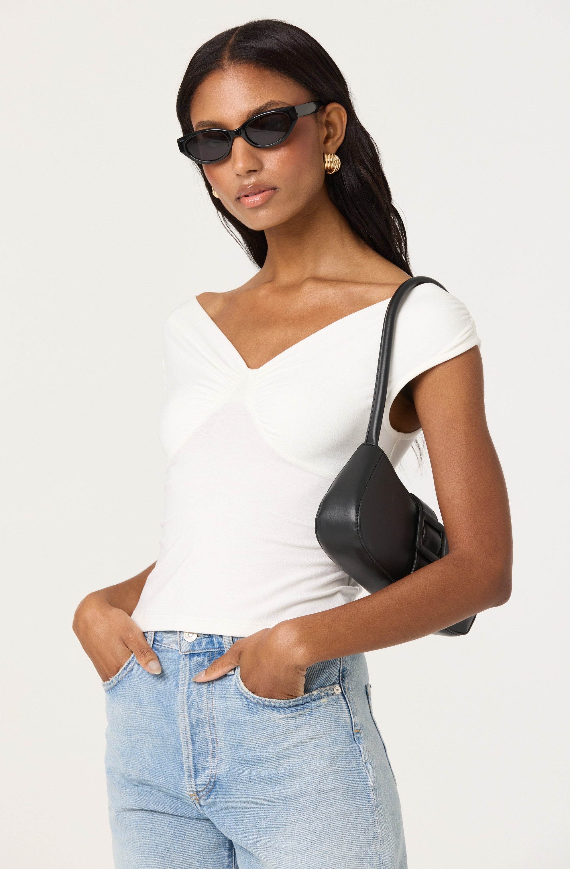 A woman is wearing a white, short-sleeved, V-neck top that features gathered detailing at the center. She is also wearing light blue high-waisted jeans. She has on black sunglasses and gold hoop earrings. She carries a black shoulder bag with a single strap over her right shoulder.