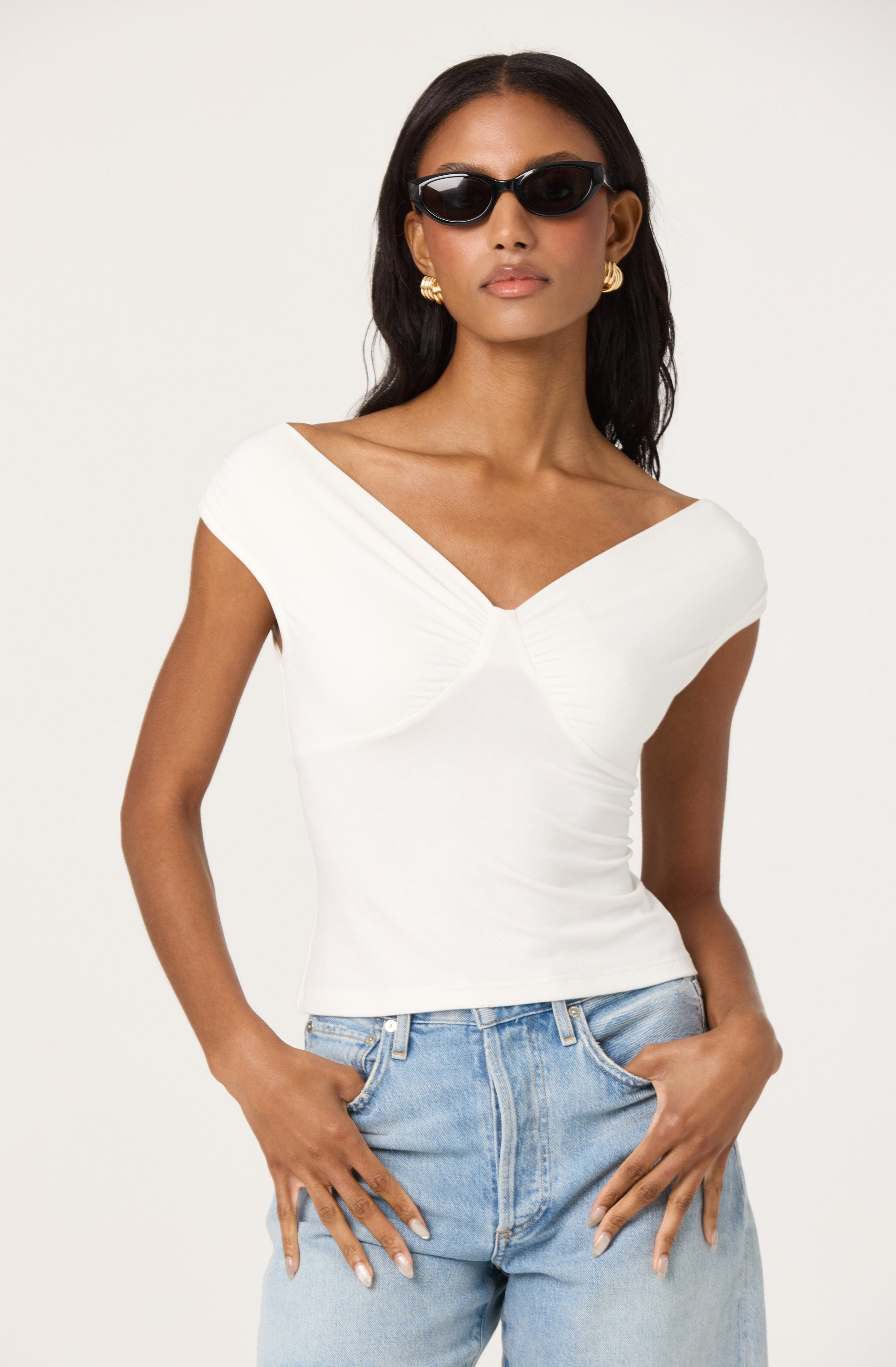 A woman is wearing a white, fitted top with a V-shaped neckline and short, gathered cap sleeves. She has paired it with light-wash, high-waisted jeans. She accessorizes with large, gold hoop earrings and dark sunglasses.