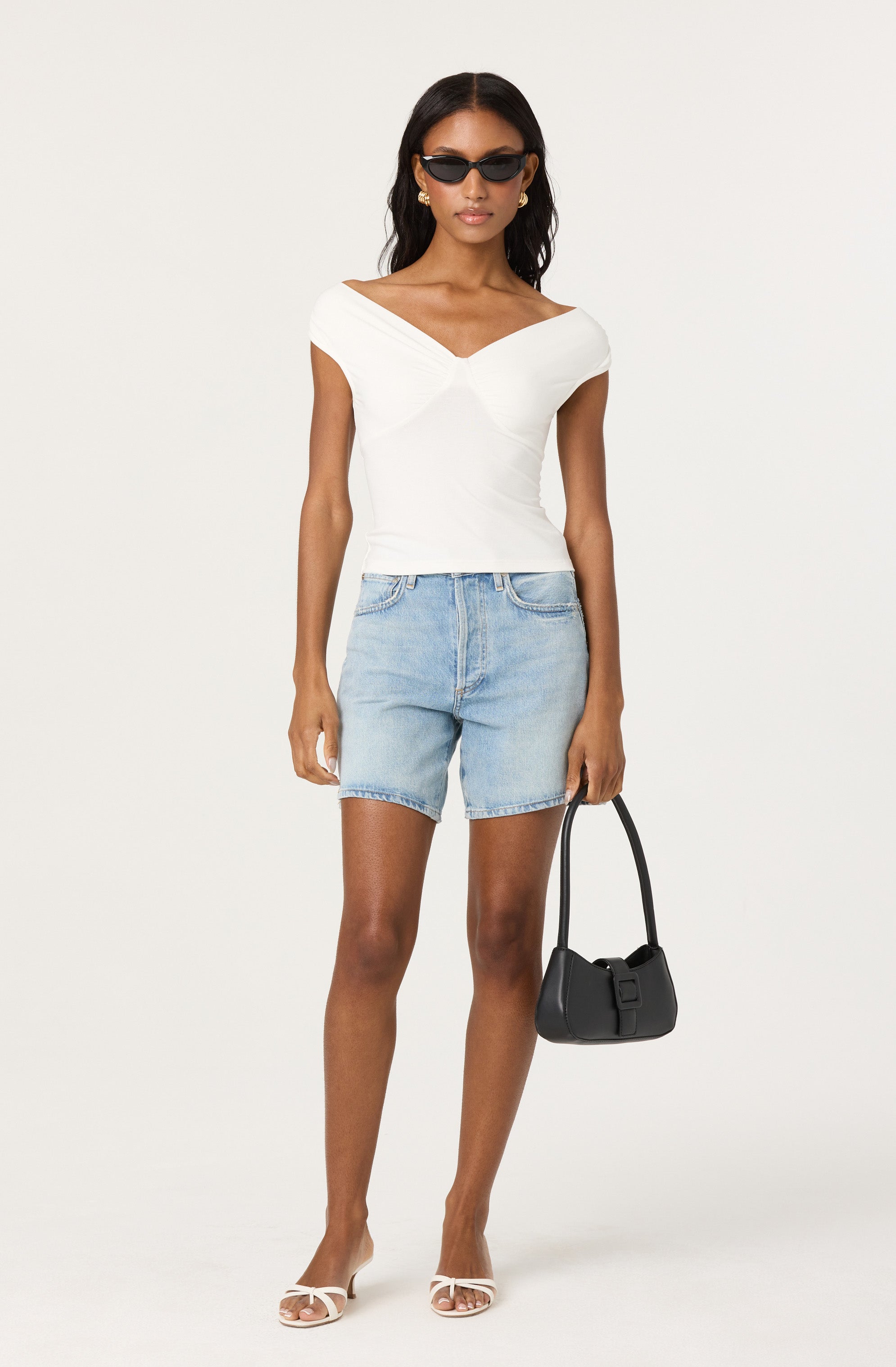 A woman is wearing a white, fitted, off-the-shoulder top with a V-shaped neckline. She has on light blue denim shorts and white high-heeled sandals with thin straps. She accessorizes with small gold earrings, dark sunglasses, and gold hoop earrings. She is holding a small black handbag with a short strap.