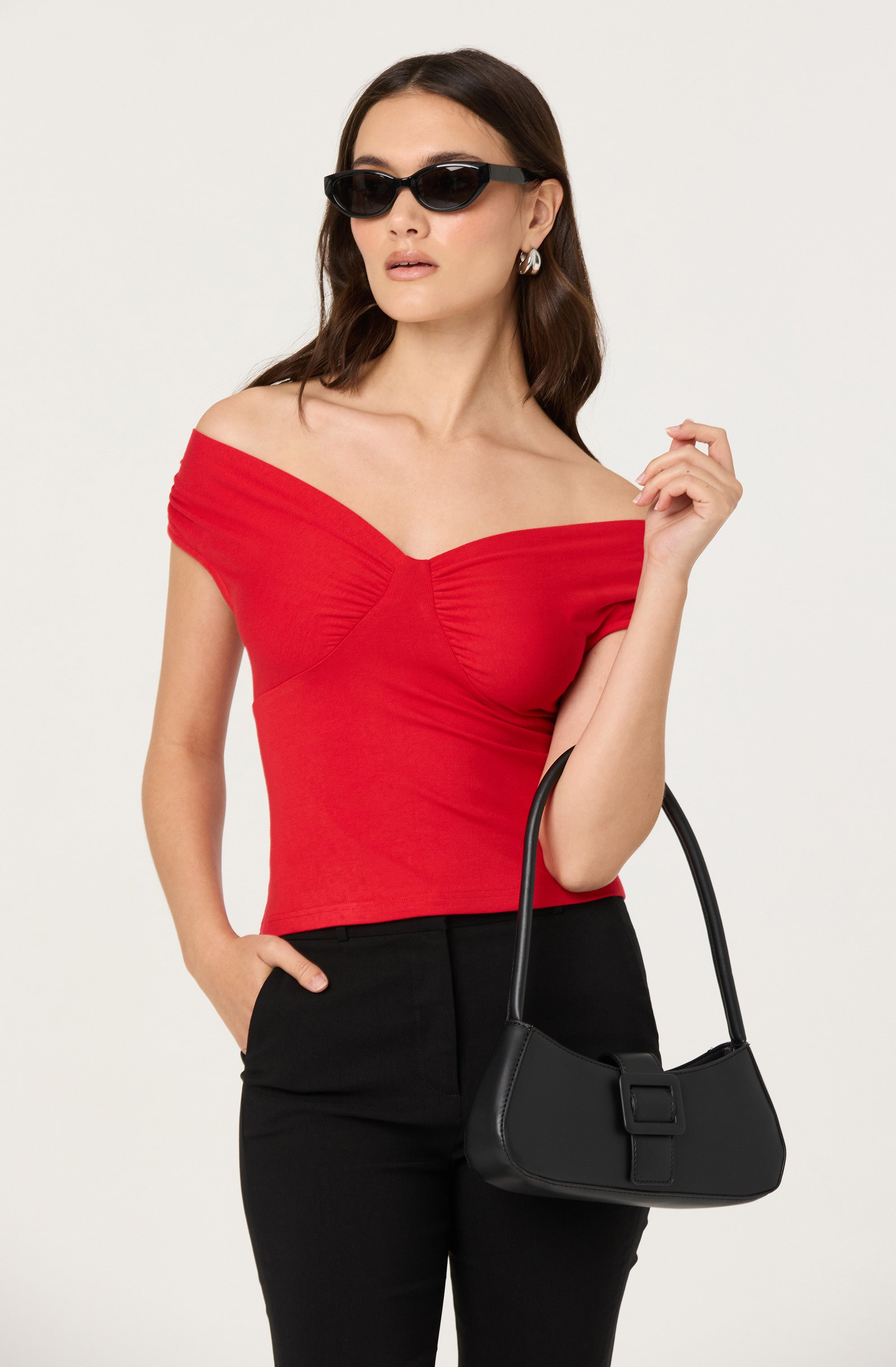 A woman is wearing a red off-the-shoulder top with gathered detailing at the bust and sleeves. She pairs it with black high-waisted pants and accessorizes with black sunglasses, silver hoop earrings, and carries a black handbag with a wide strap.