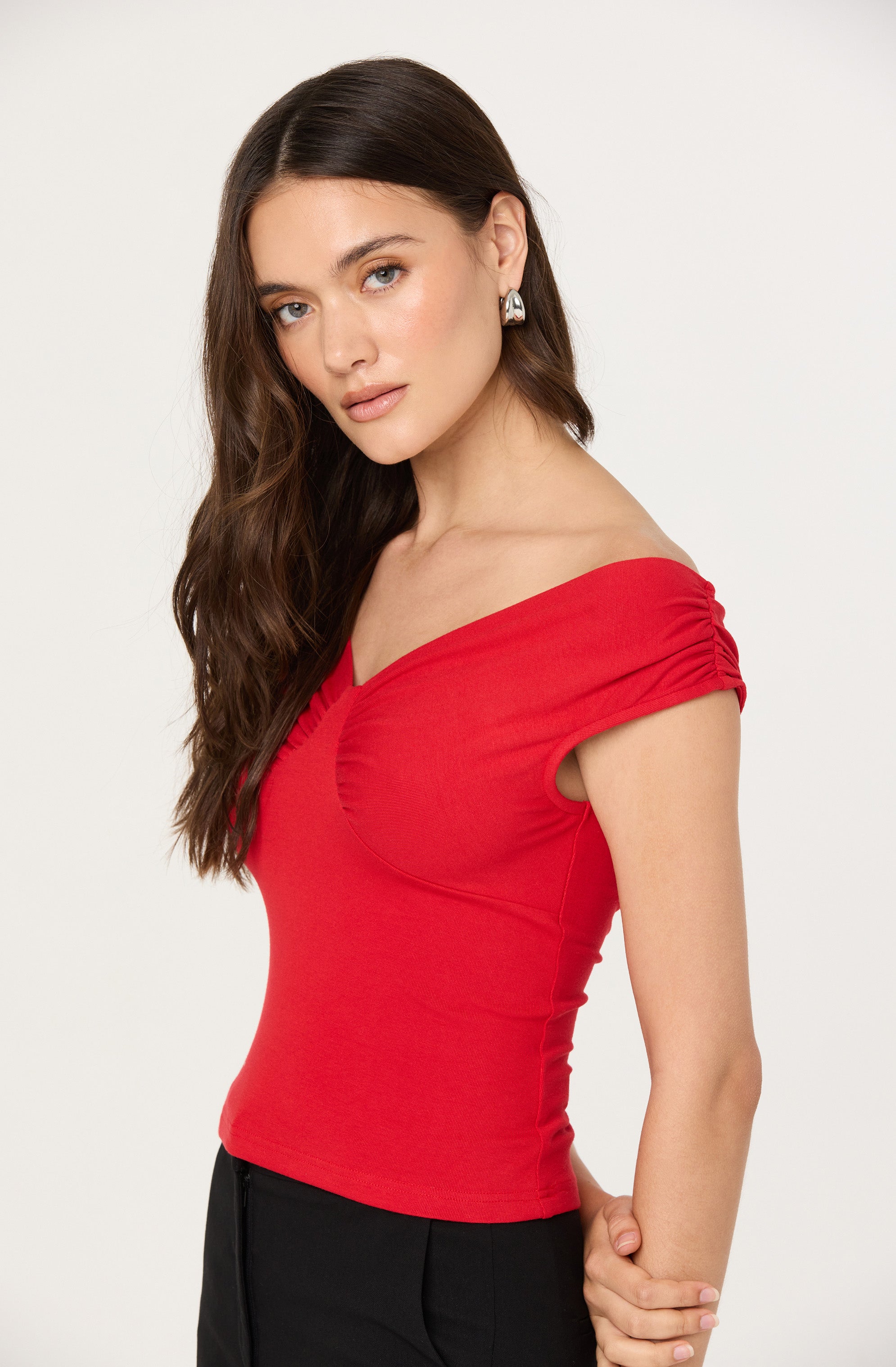 A woman is wearing a fitted red top with ruched detailing on the short, capped sleeves and along the neckline. She has long, wavy brown hair, accessorized with large, silver hoop earrings.