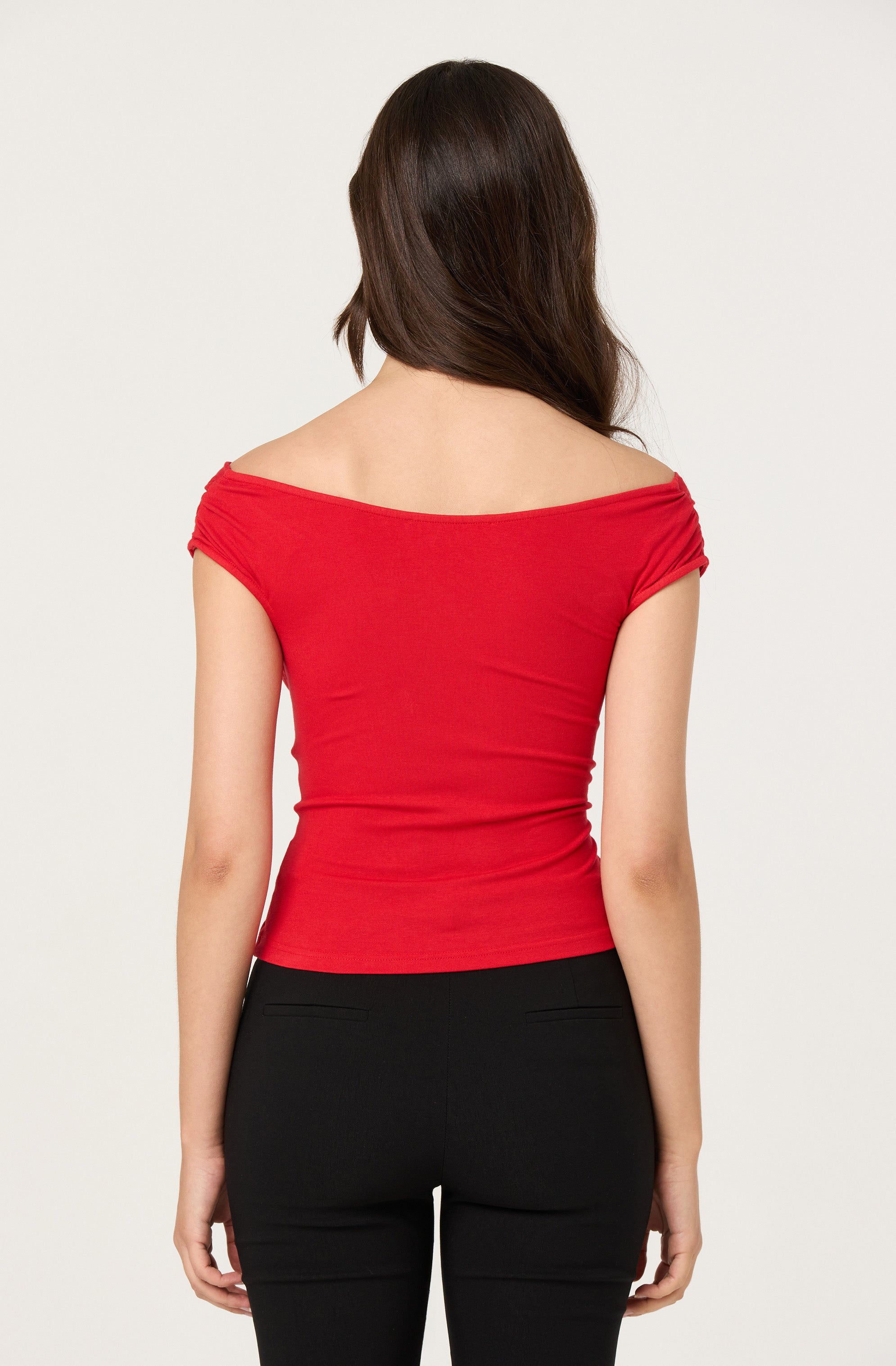 A woman is wearing a fitted, off-the-shoulder, short-sleeve top in bright red. The top has gathered fabric at the shoulders and a wide neckline that rests on her upper arms. She is also wearing black pants that are snug and have back pockets. The background is plain and light-colored.