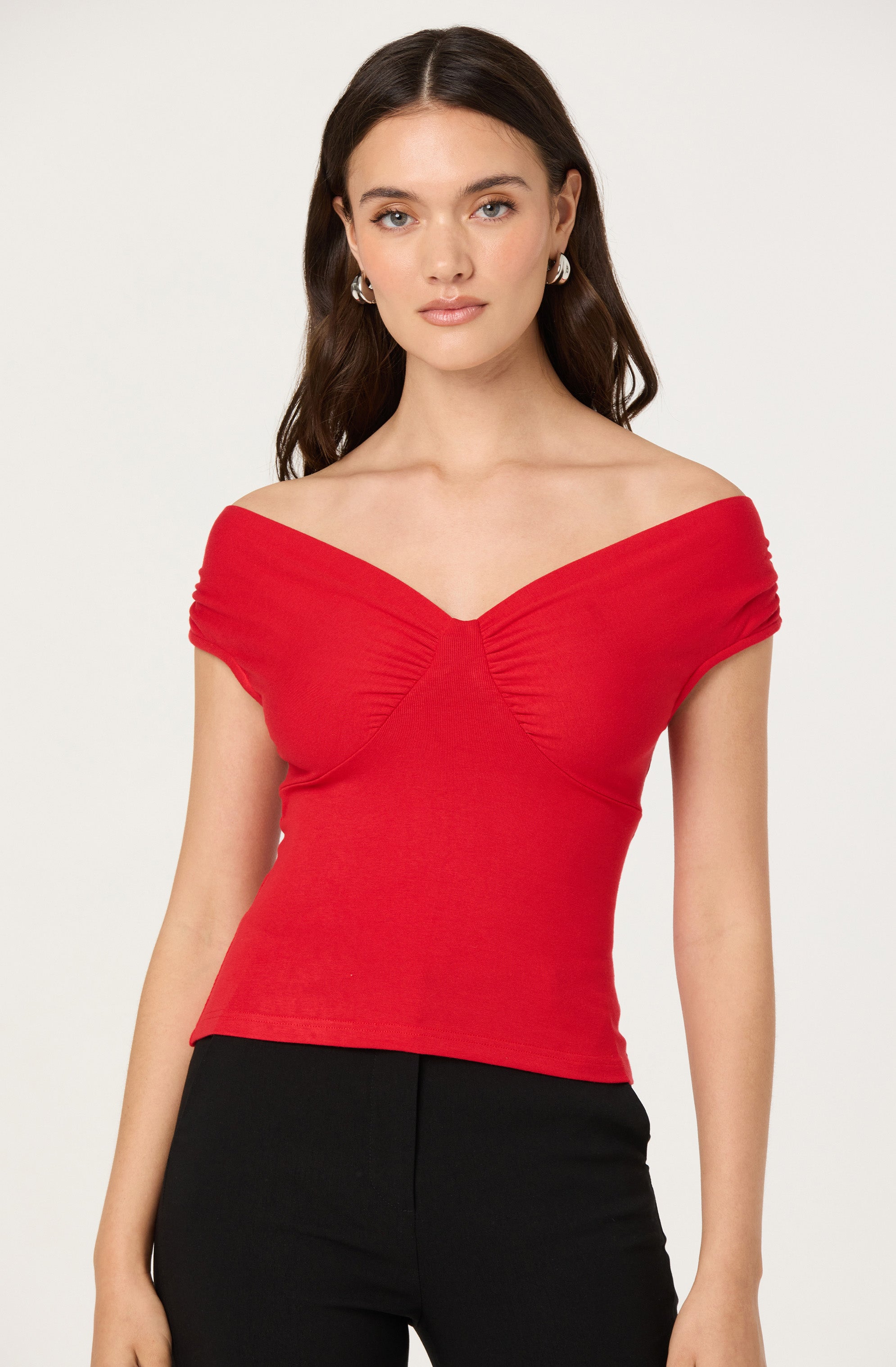 A woman is wearing an off-the-shoulder red short-sleeve top with ruched detailing at the bust and fitted black pants.